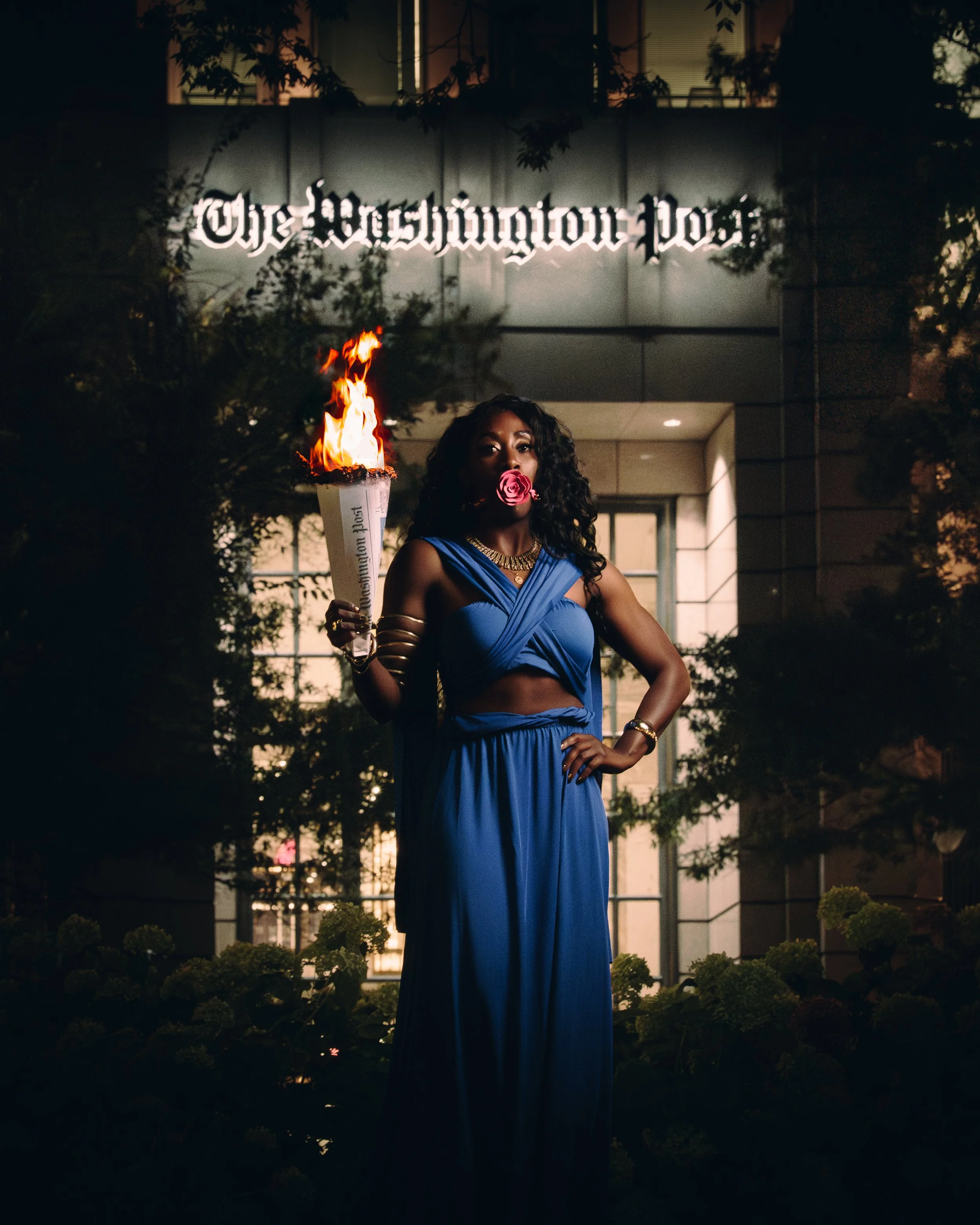 A woman in a blue dress holding a burning torch with the words 'The Washington Post' written on it, standing in front of a building with a sign 'The Washington Post' illuminated above her at night.