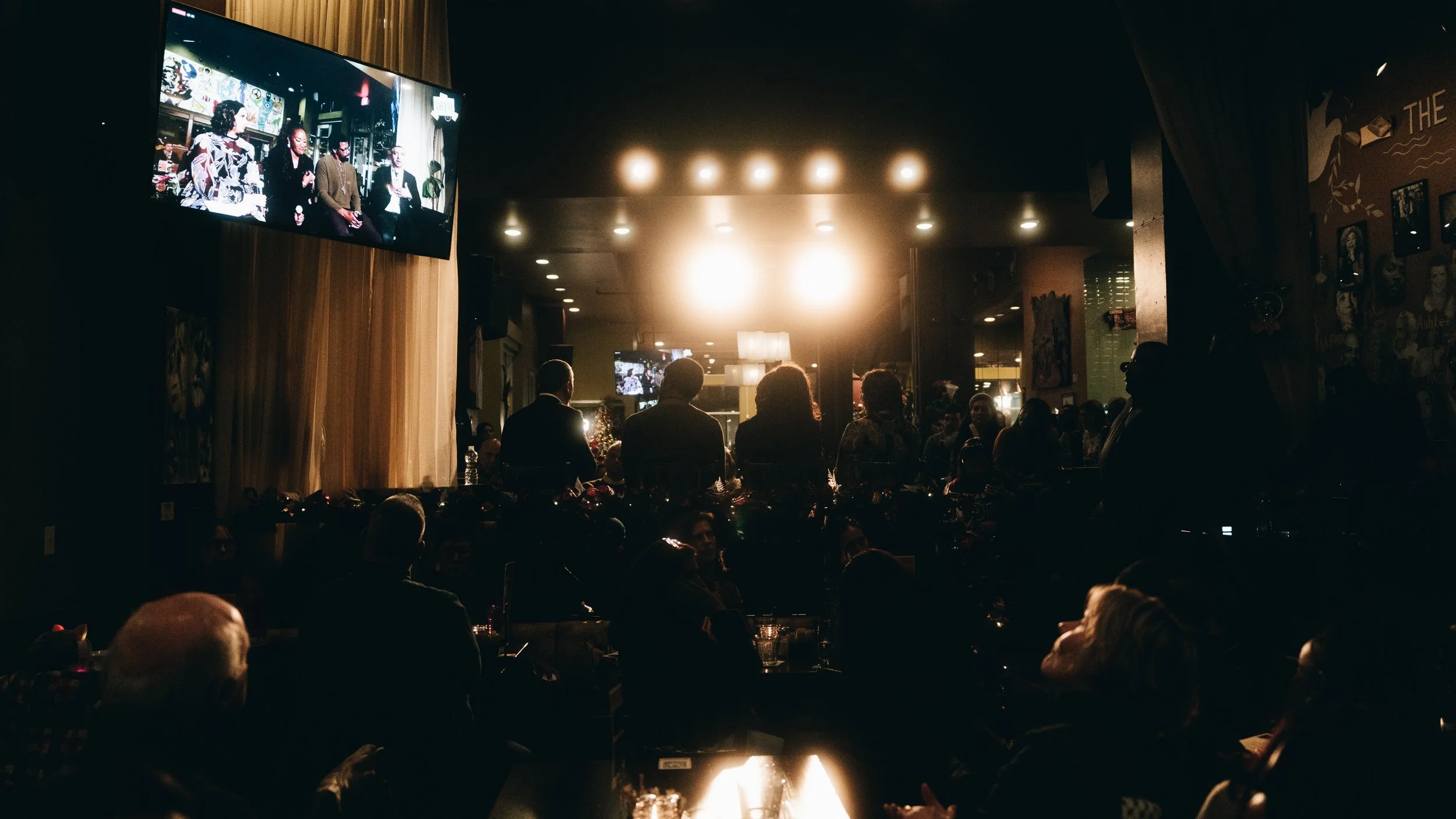 People watching a TV screen in a darkened bar or restaurant, with bright lights and silhouettes of patrons visible.