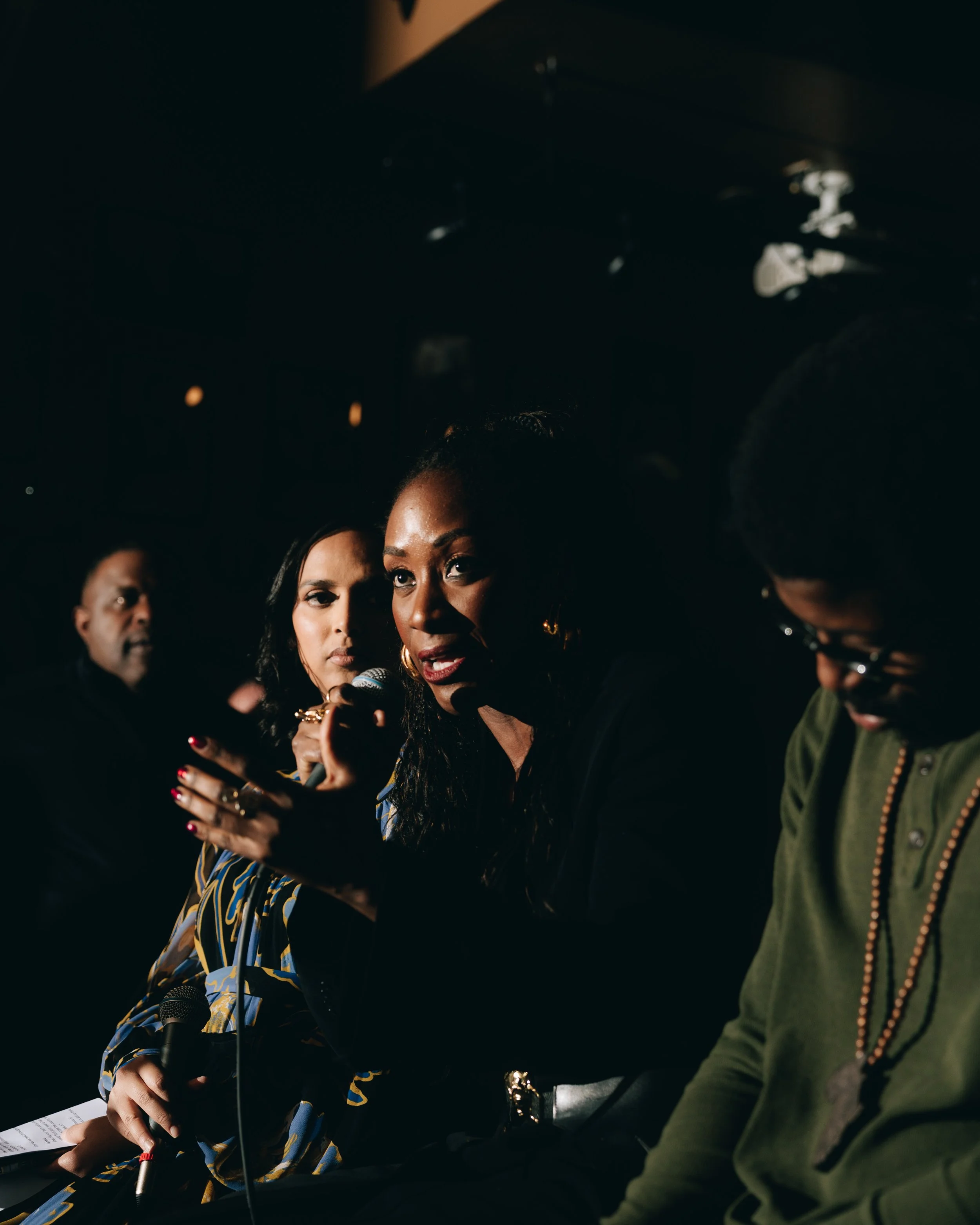 A woman holding a microphone, speaking at a panel discussion. Two other people are seated beside her, one on each side, partially visible in the dark background.