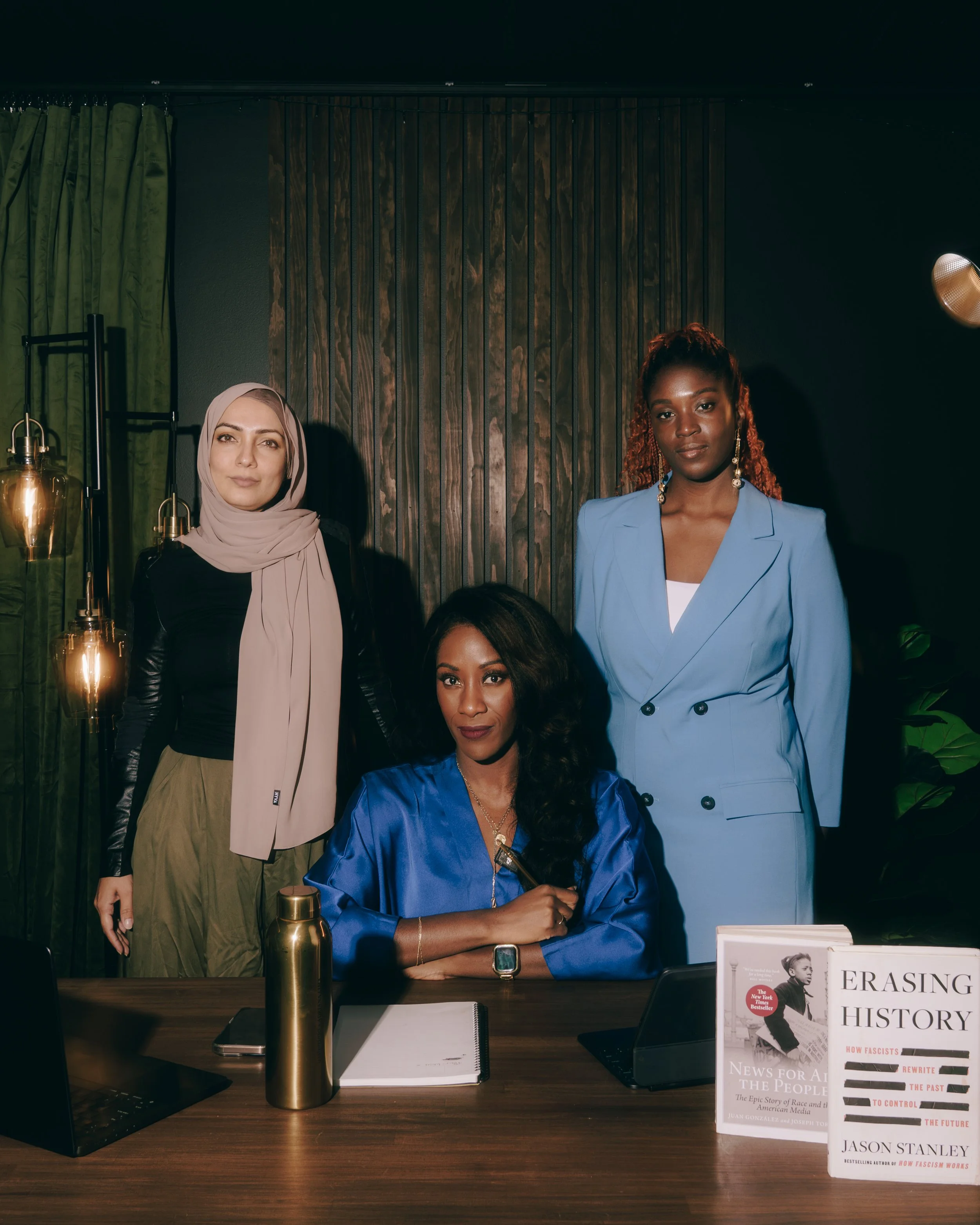 Three women in a professional setting, with one sitting at a desk and two standing behind her. The sitting woman wears a blue satin jacket, the standing women wear a light blue suit and an outfit with a hijab, and there are books and electronic devic