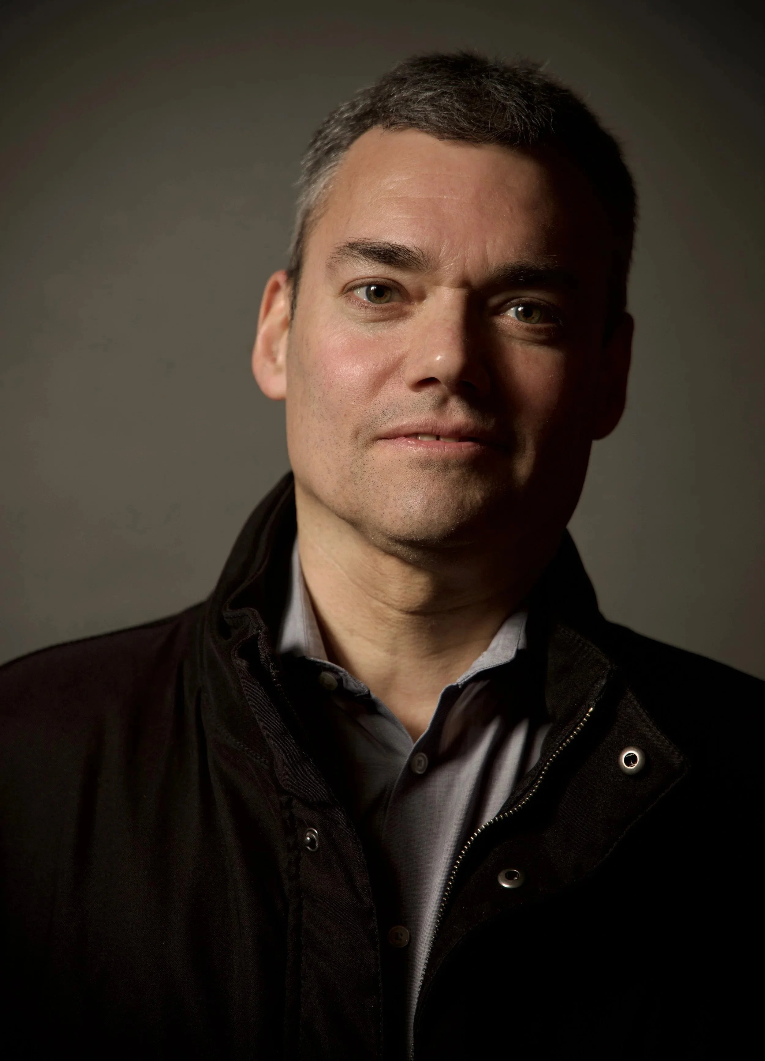 A man with short dark hair, wearing a dark jacket over a light collared shirt, posing against a dark background.