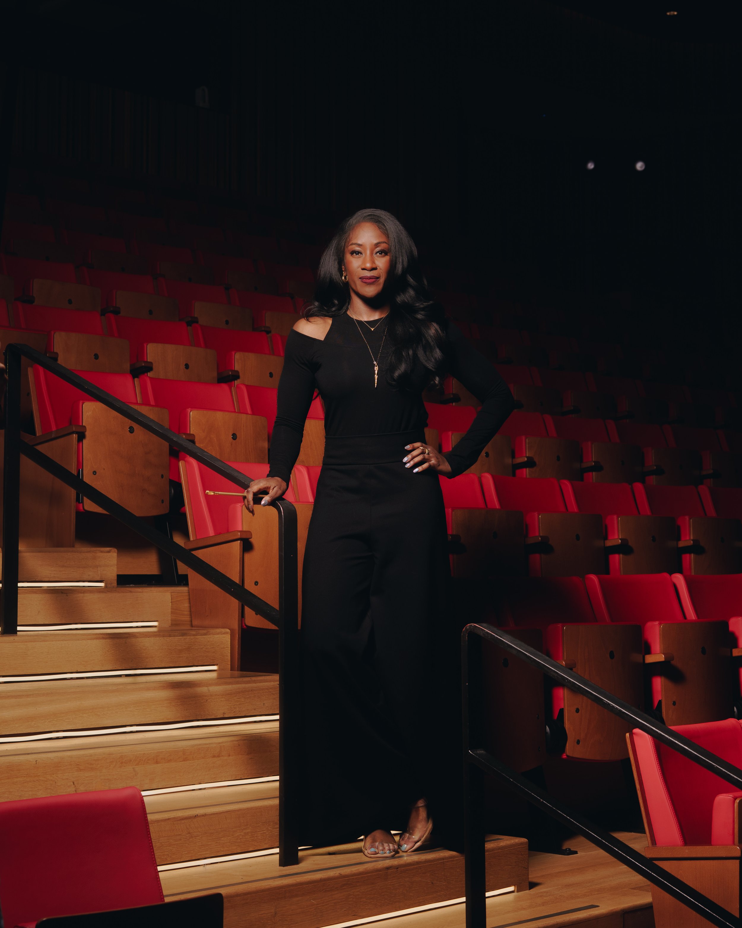 A woman with long black hair wearing a black off-the-shoulder dress and high heels stands on a wooden staircase in an auditorium with red chairs.