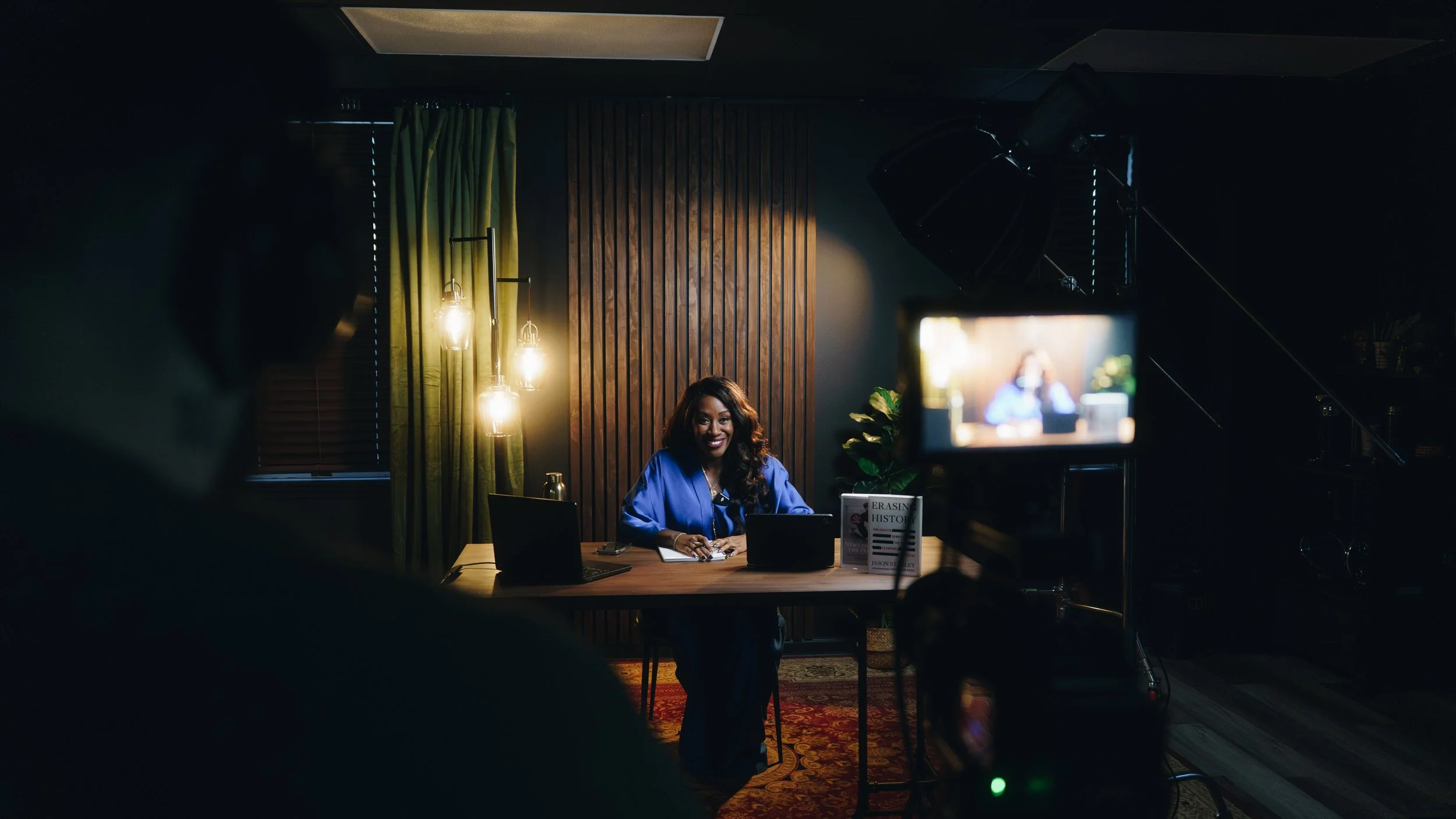 A woman sitting at a desk in a dimly lit room, being filmed during a video recording. She is smiling, wearing a blue dress, and surrounded by laptops, with a camera and lighting equipment directed at her.