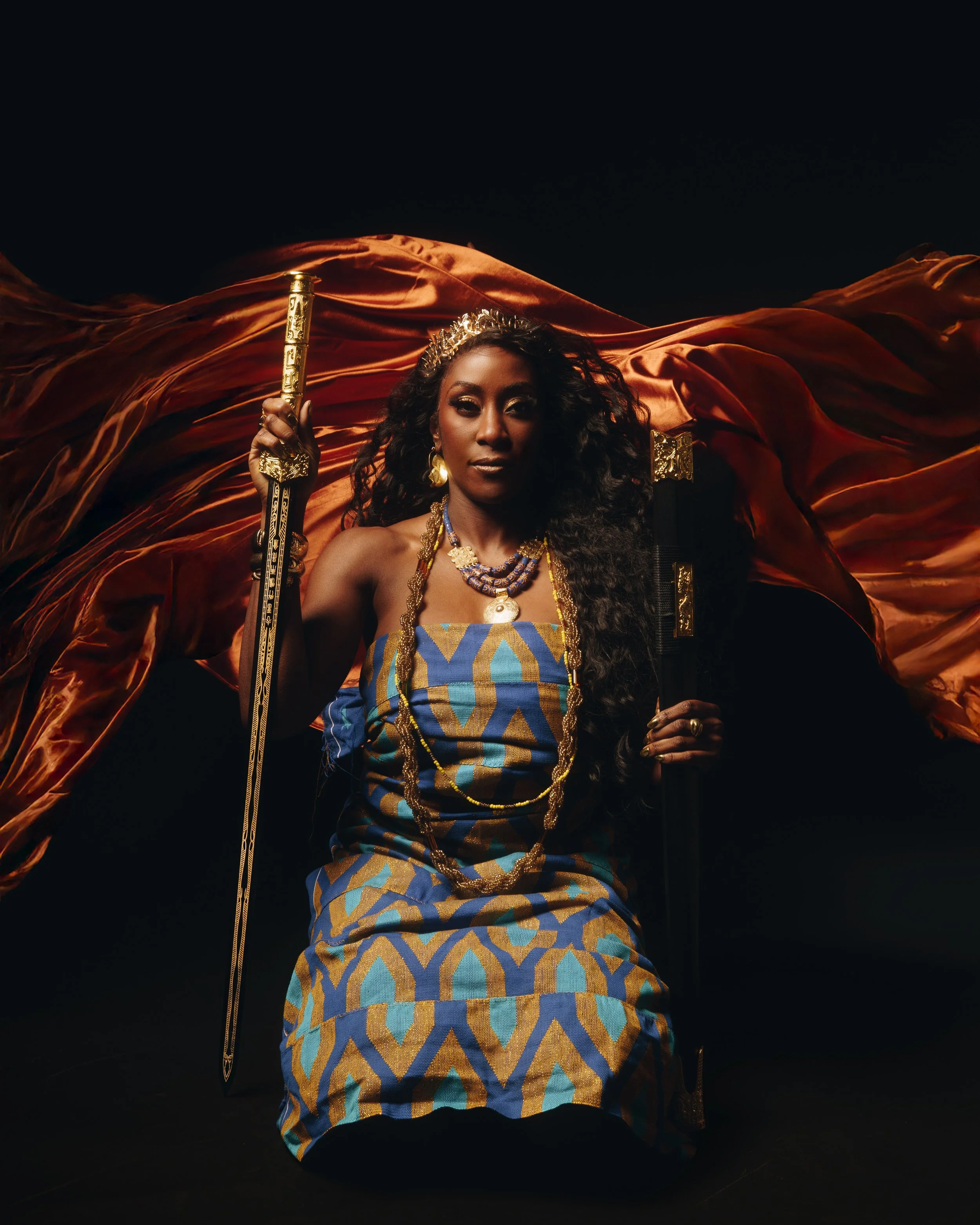 A woman dressed in traditional African attire holding a staff, wearing jewelry and a crown, with a flowing orange fabric in the background.