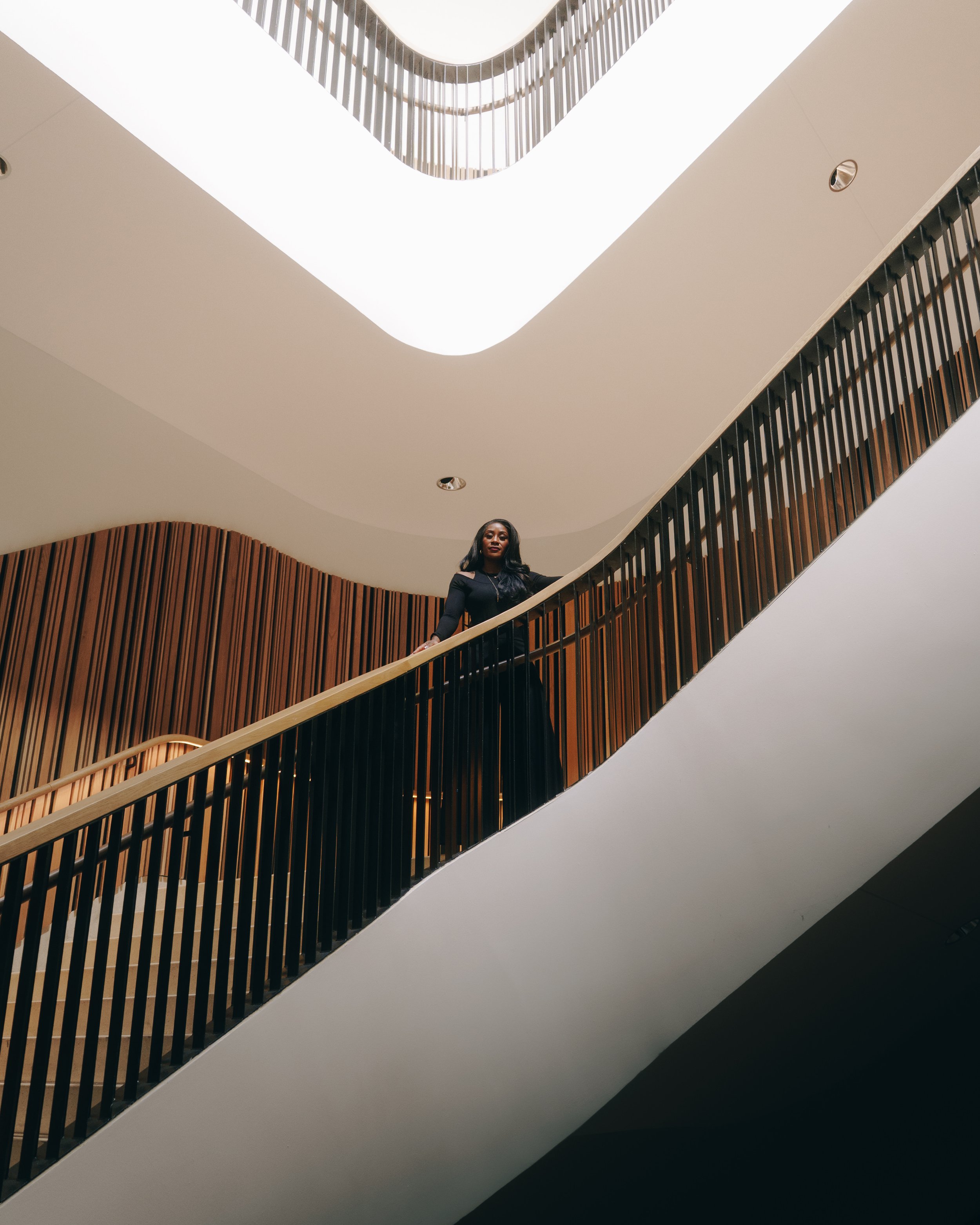 A woman in a black dress standing on a modern staircase with wooden railings, looking down from a lower level.
