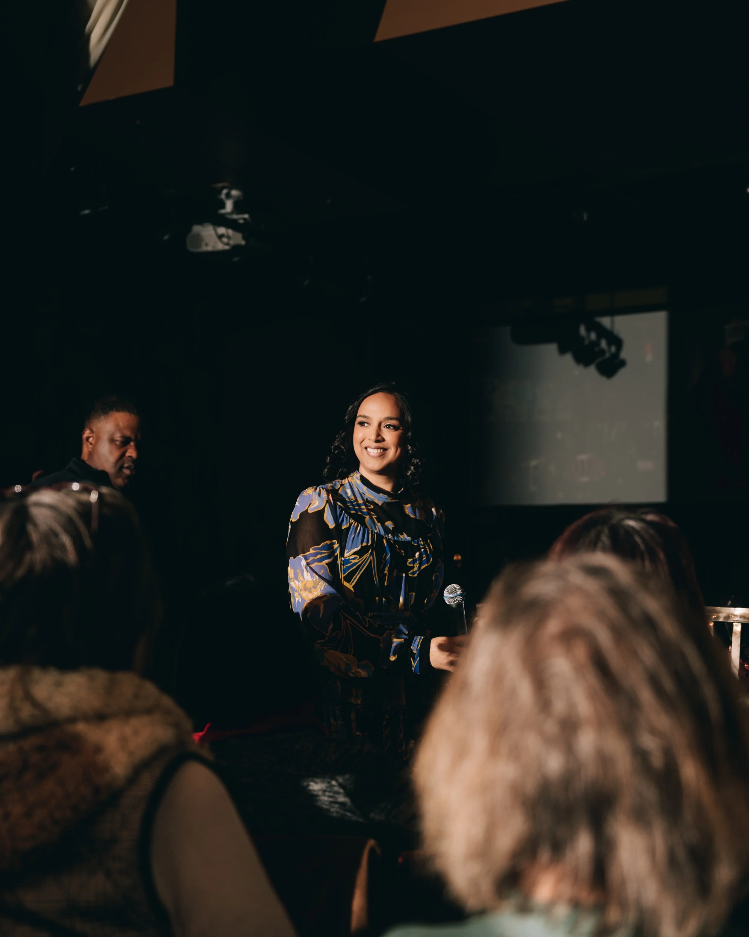 A woman smiling and holding a microphone on stage during a presentation or speech, with an audience in front of her in a dimly lit room.