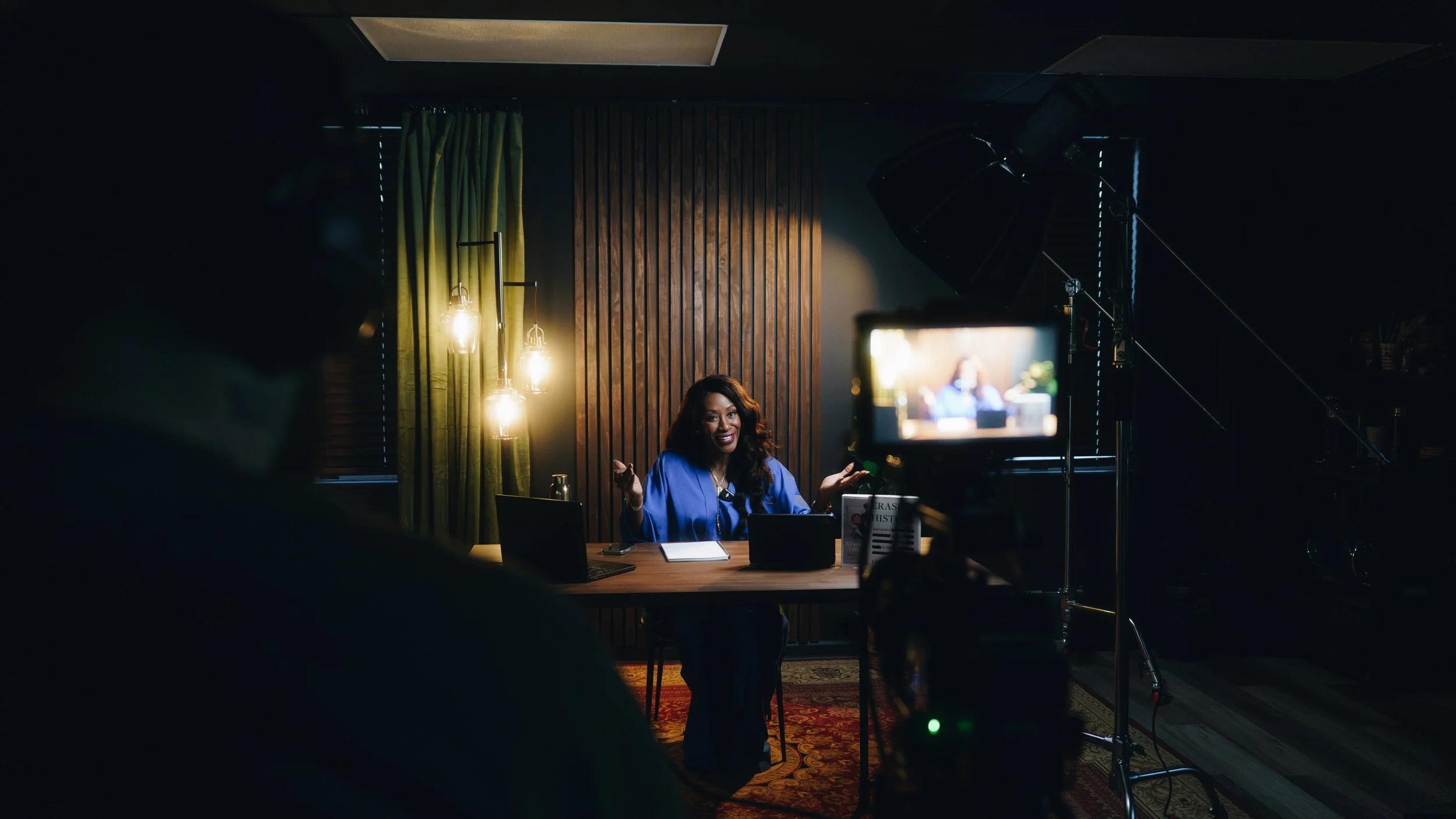 A woman in a blue blazer is sitting at a desk during a video recording or interview, with a camera filming her, in a dimly lit room with wooden paneling, yellow curtains, and pendant lights.