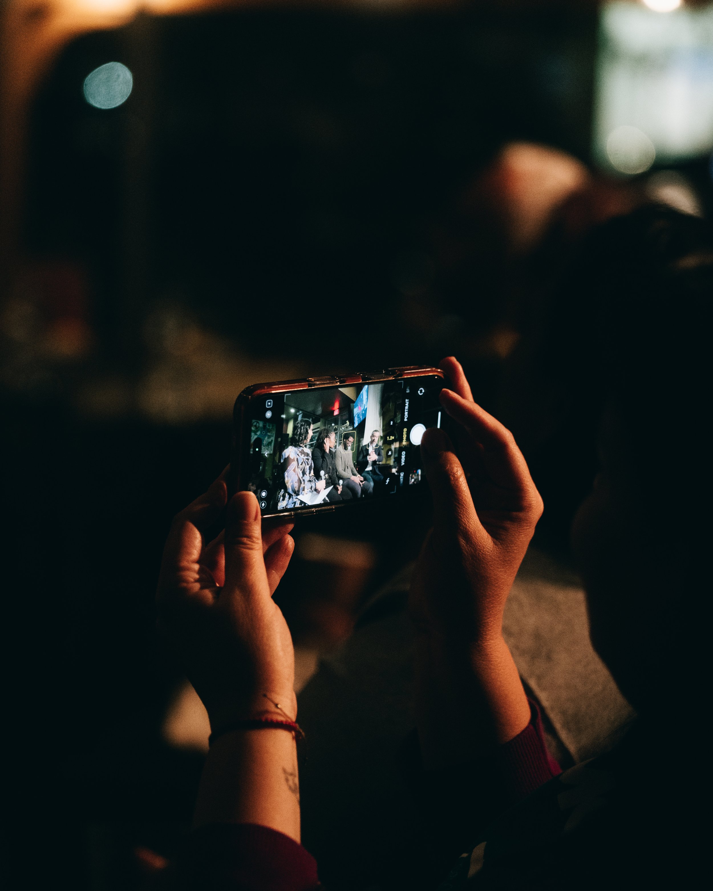 Person taking a photo or recording a video of a panel discussion or interview on a smartphone in a dimly lit setting.