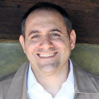 Close-up of a smiling man with short dark hair, wearing a light-colored shirt and jacket, against a wooden background.
