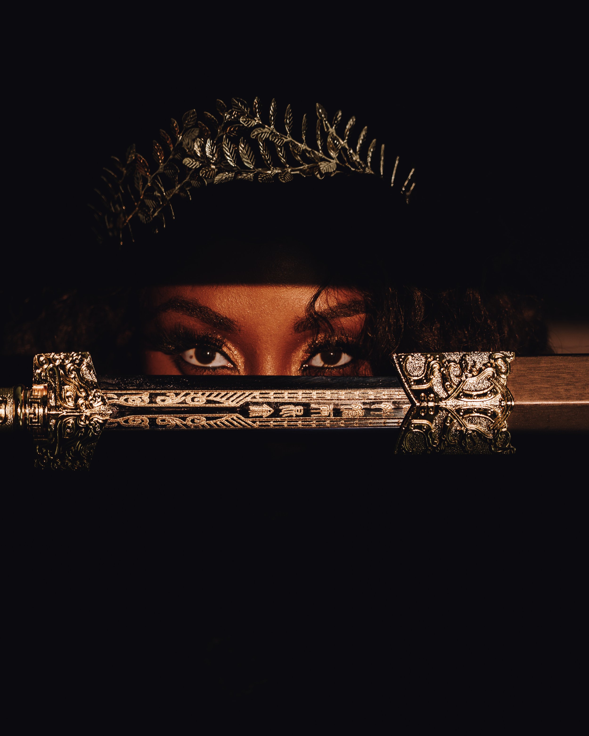 A woman with dark makeup and piercing eyes wearing a jeweled crown, holding a shiny, ornate sword horizontally in front of her face in a dark setting.