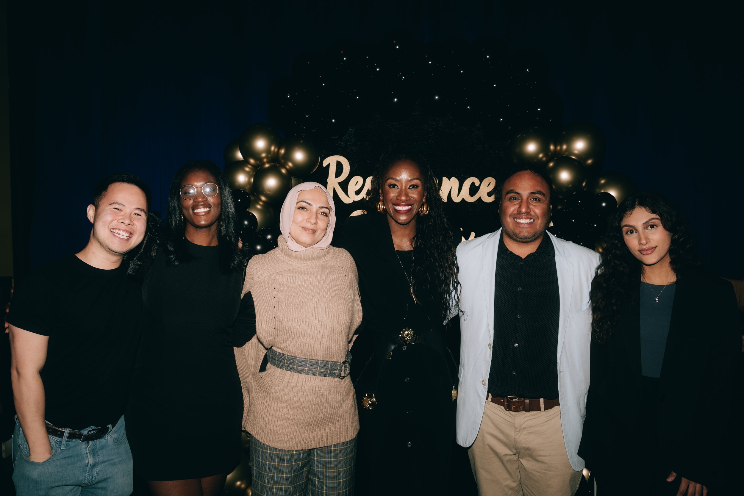 Group of six people smiling in front of a black backdrop with gold balloons