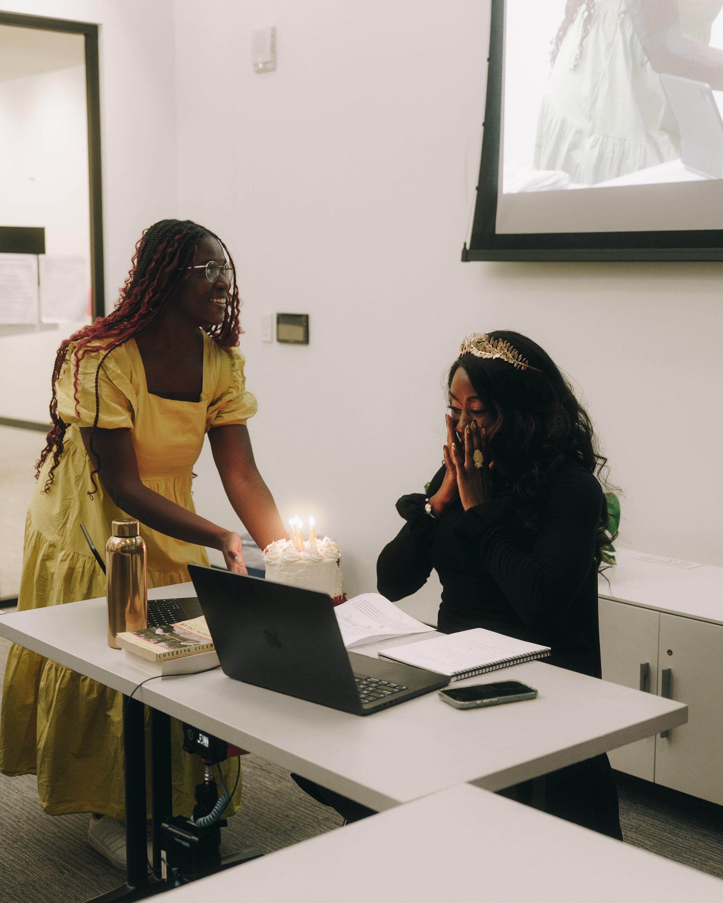A woman wearing a yellow dress is standing and holding a birthday cake with lit candles, smiling. Another woman dressed in black with a gold crown is sitting at a table with her hands covering her face, appearing surprised and happy.