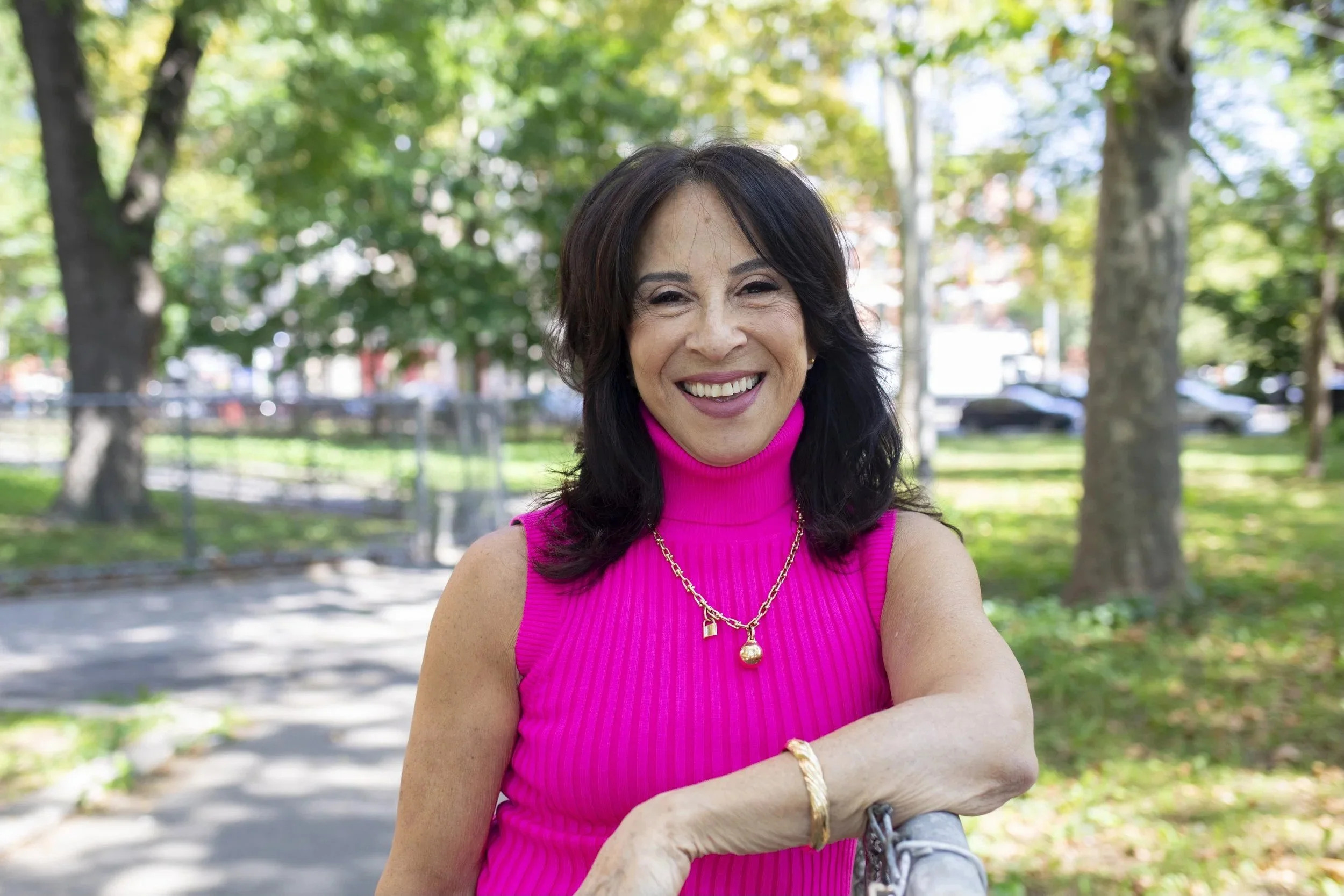 Woman in a bright pink sleeveless turtleneck top smiling outdoors in a park with trees and grass.