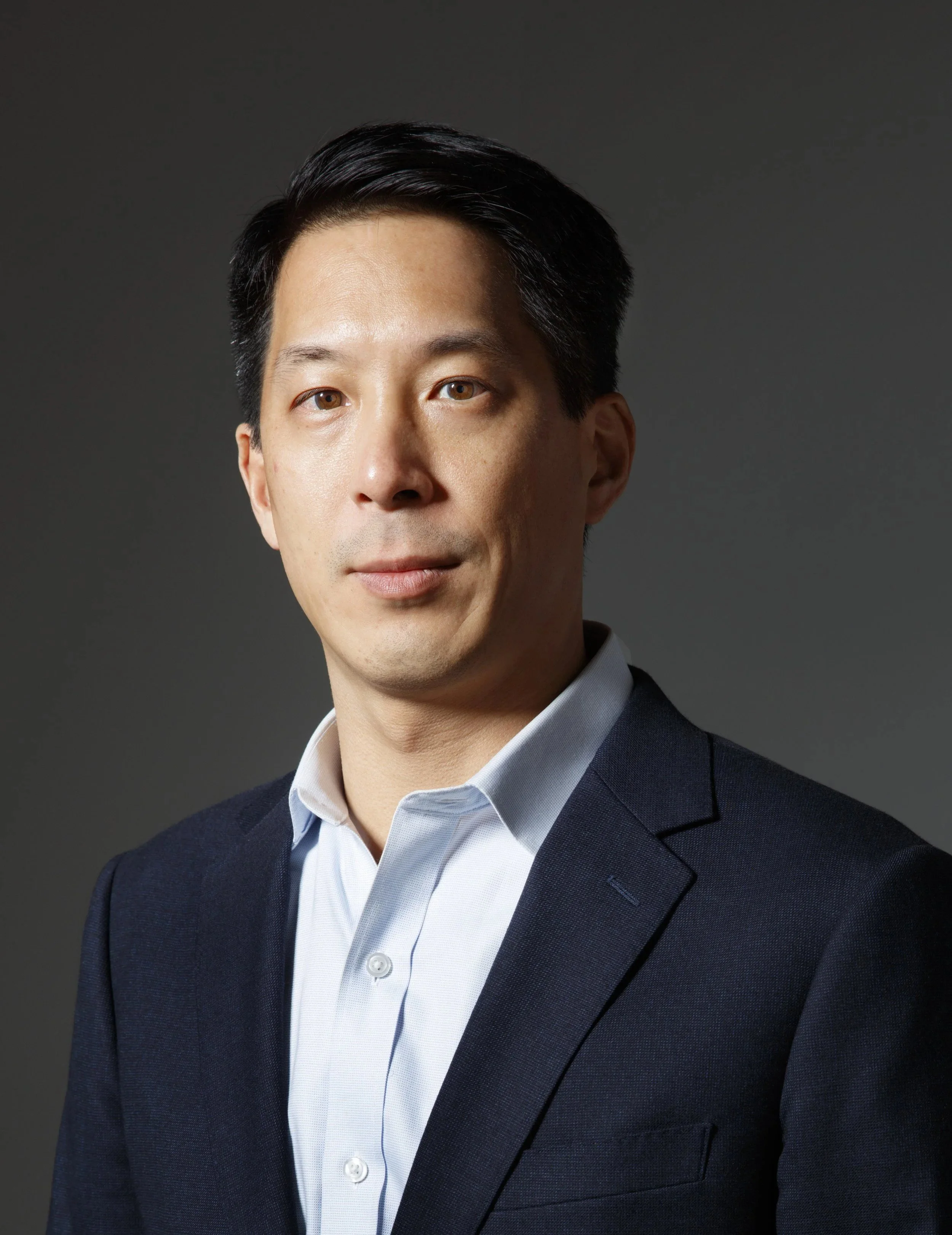 Professional portrait of man with black hair, wearing a dark suit and white dress shirt, against a gray background.