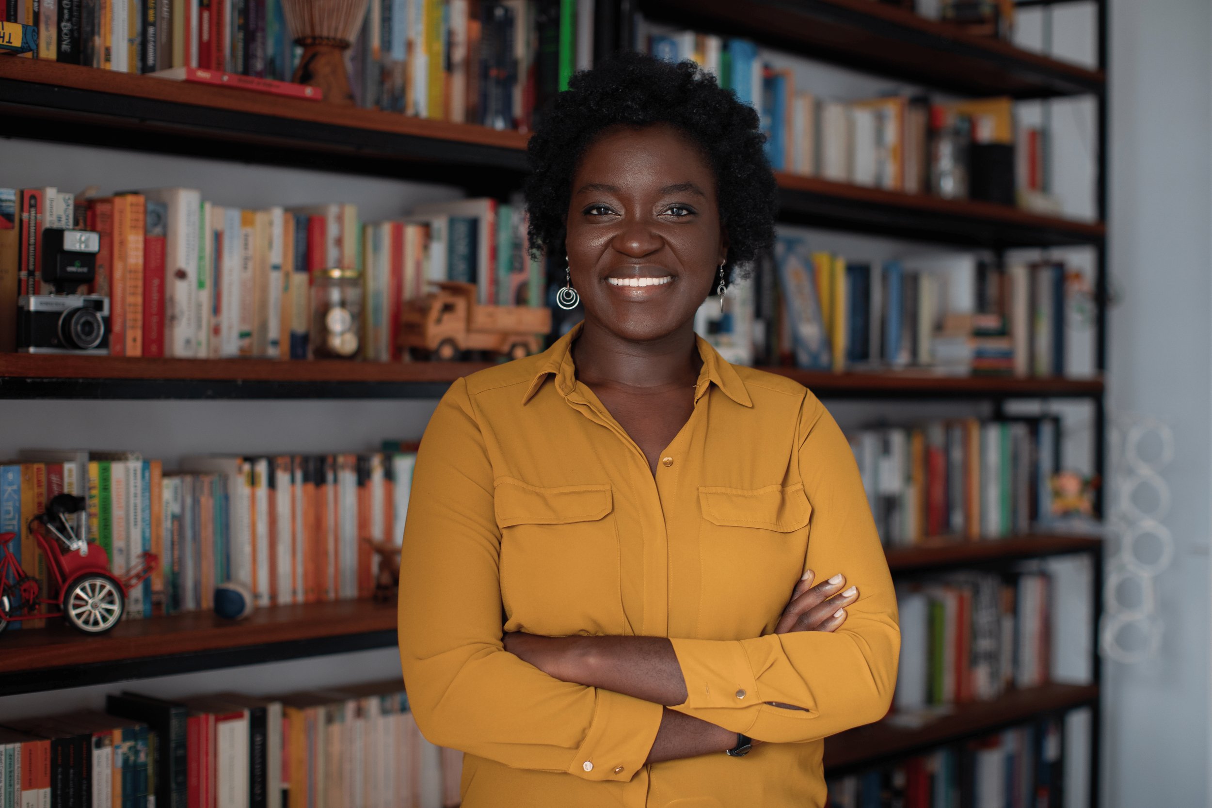 A confident woman with natural hair in a mustard yellow blouse standing in front of a bookshelf filled with books, a camera, and decorative items.