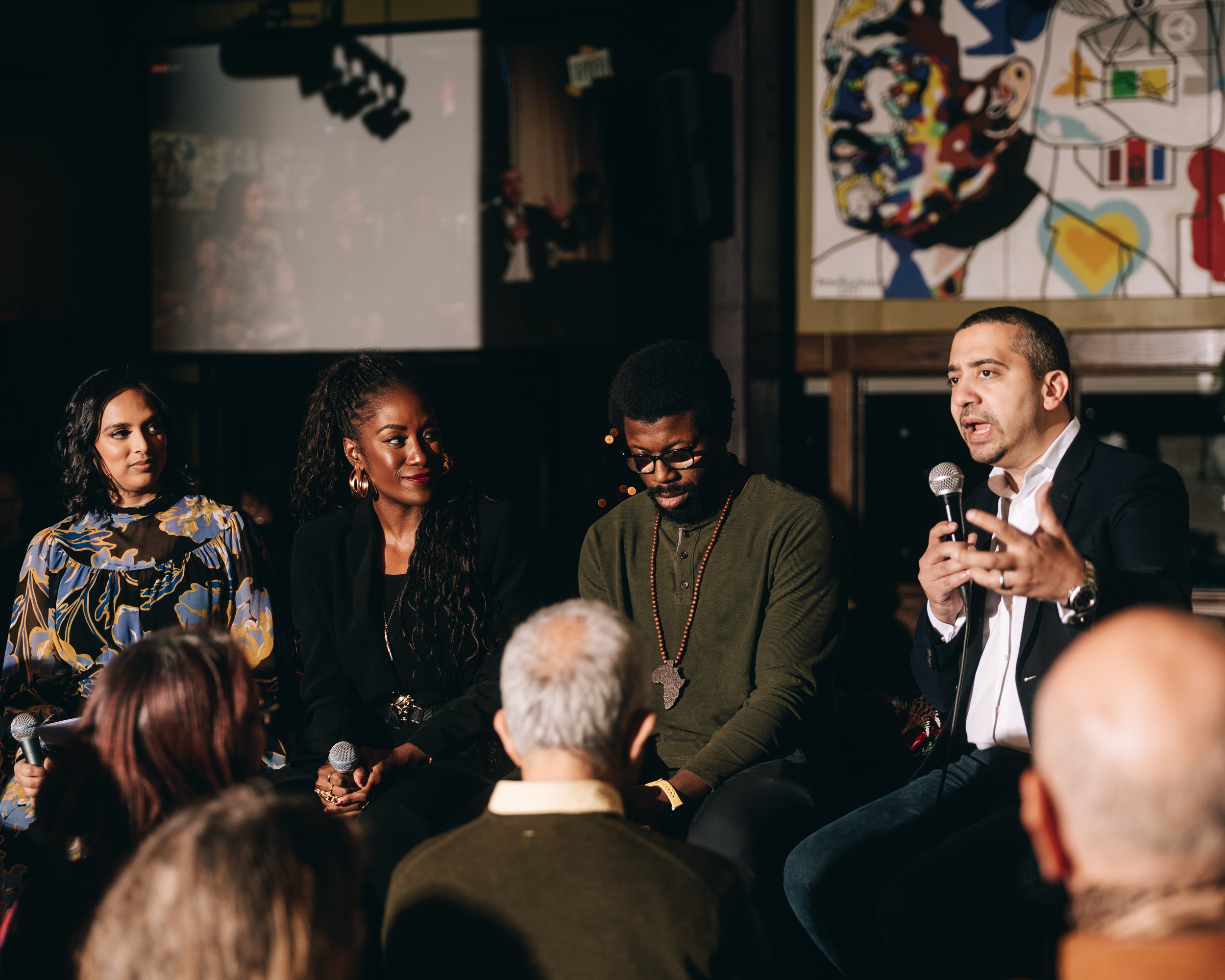 A diverse group of five people participating in a panel discussion, with one man speaking into a microphone, in a dimly lit indoor setting with artwork on the wall and audience members in the foreground.