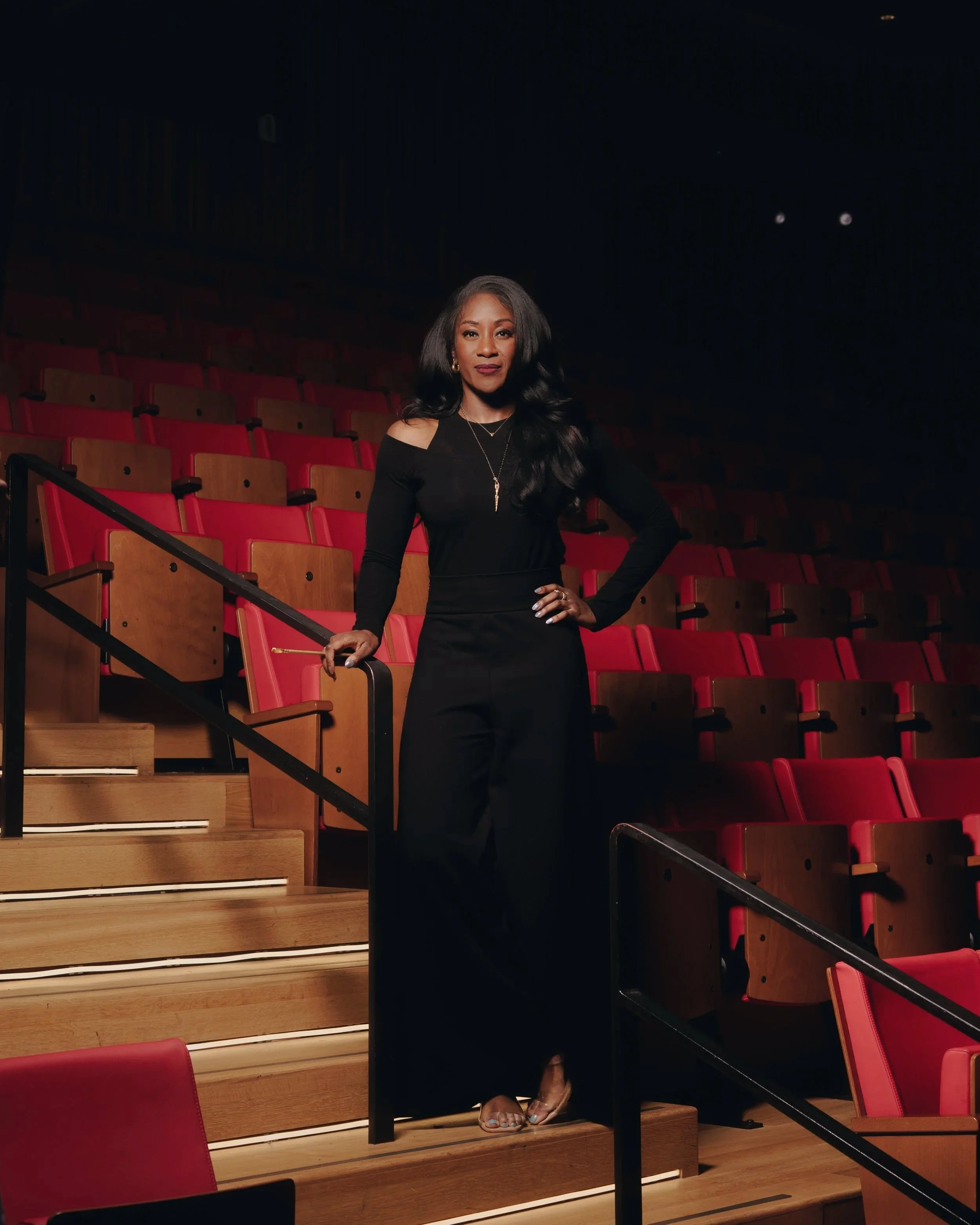 A woman dressed in black standing on theater stairs with red seats in the background.
