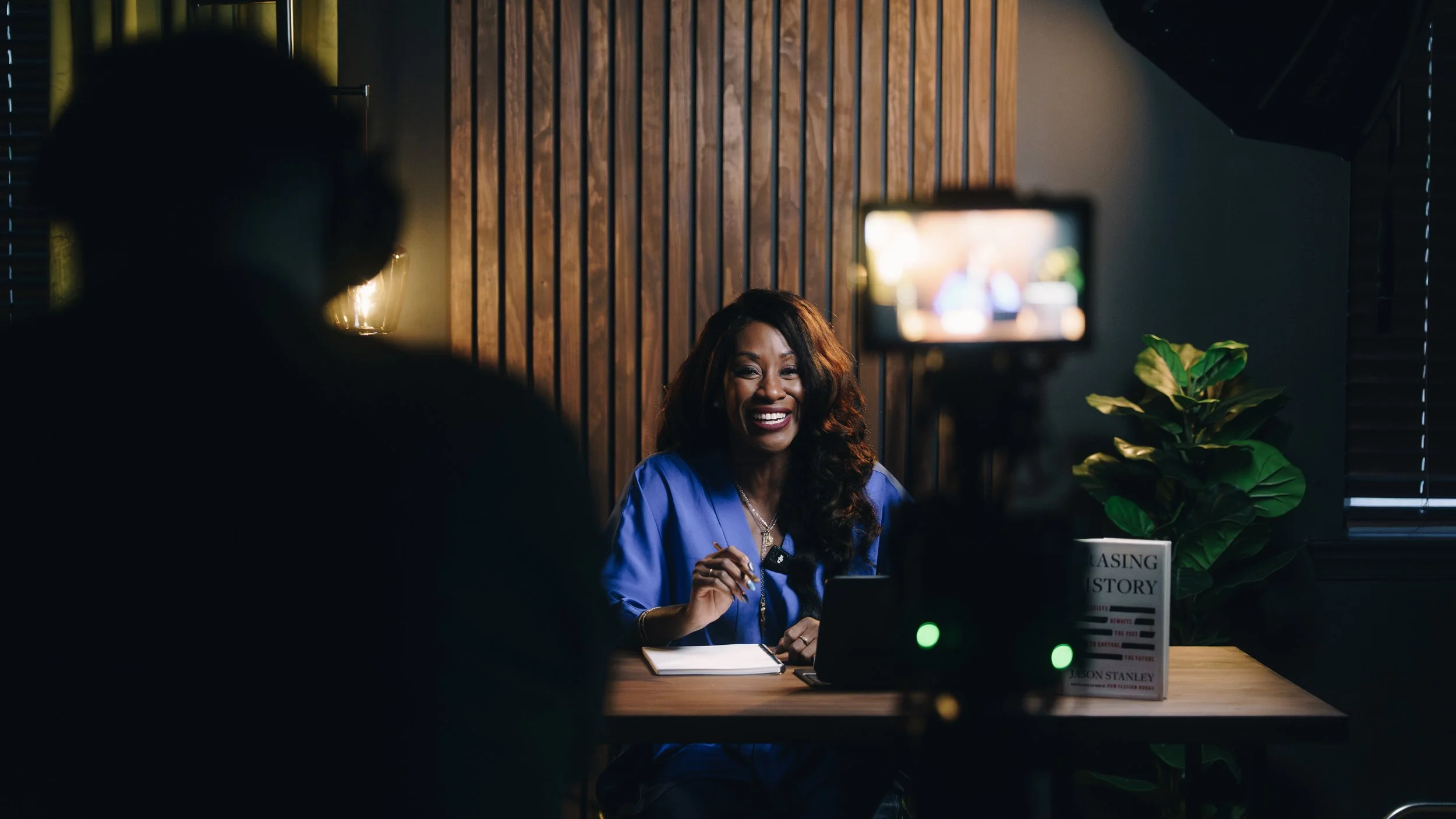 A woman is recording a video of herself, sitting at a table in a dimly lit room with wooden paneling. She is smiling and holding a pen, with a notebook open in front of her. A camera on a tripod is in the foreground, focused on her, and a large plant is visible in the background.