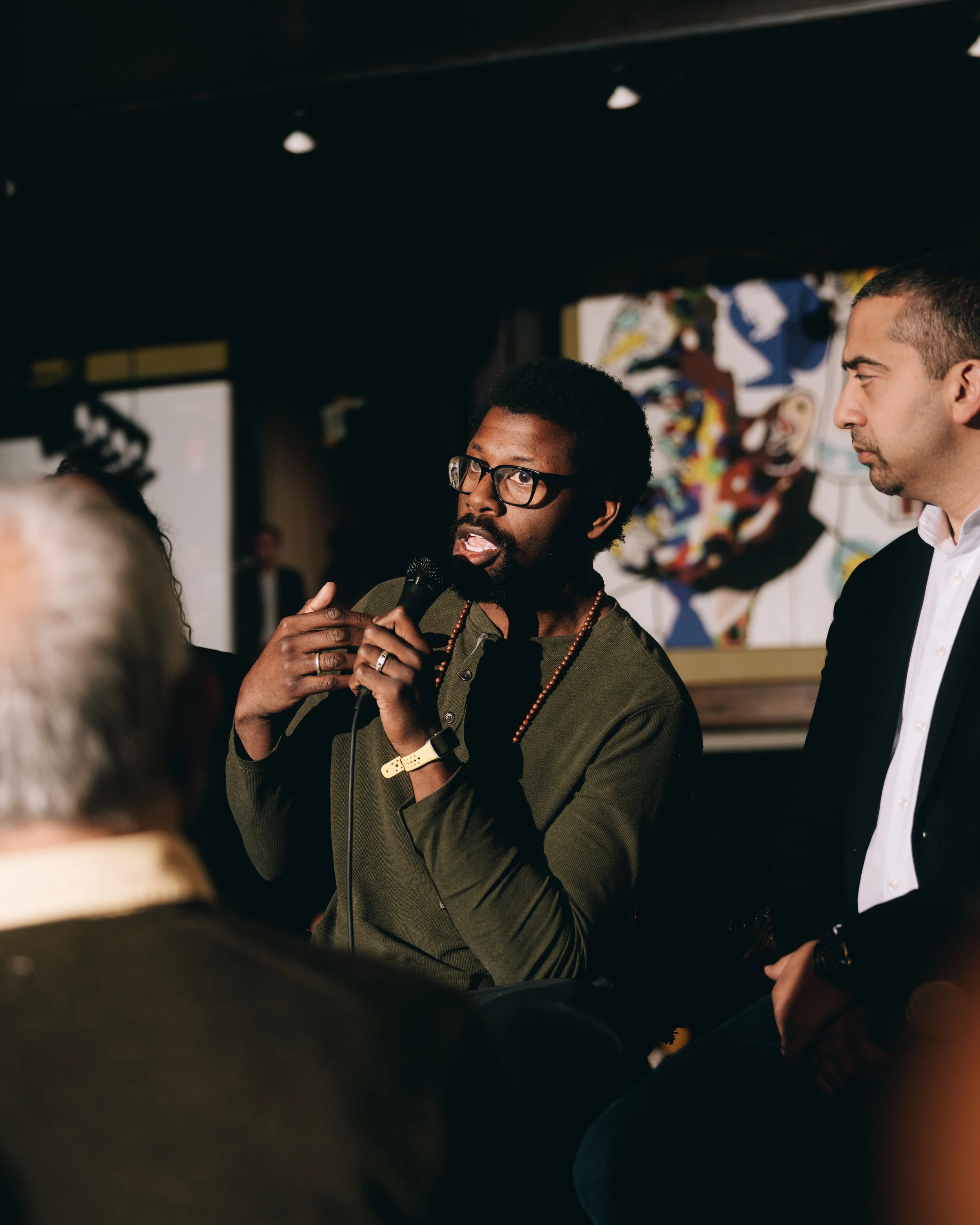 A man with glasses, a beard, and curly hair speaking into a microphone at an event, with a man in a suit listening nearby, and a colorful abstract painting in the background.