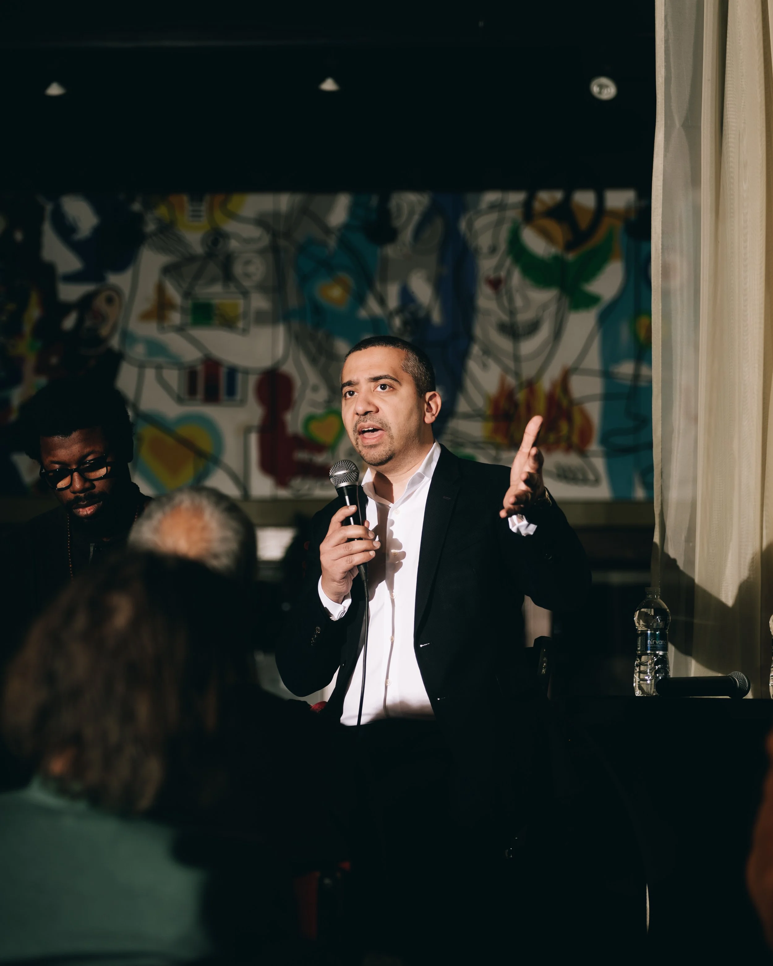 Man in black suit speaking into a microphone at a public event with an audience.