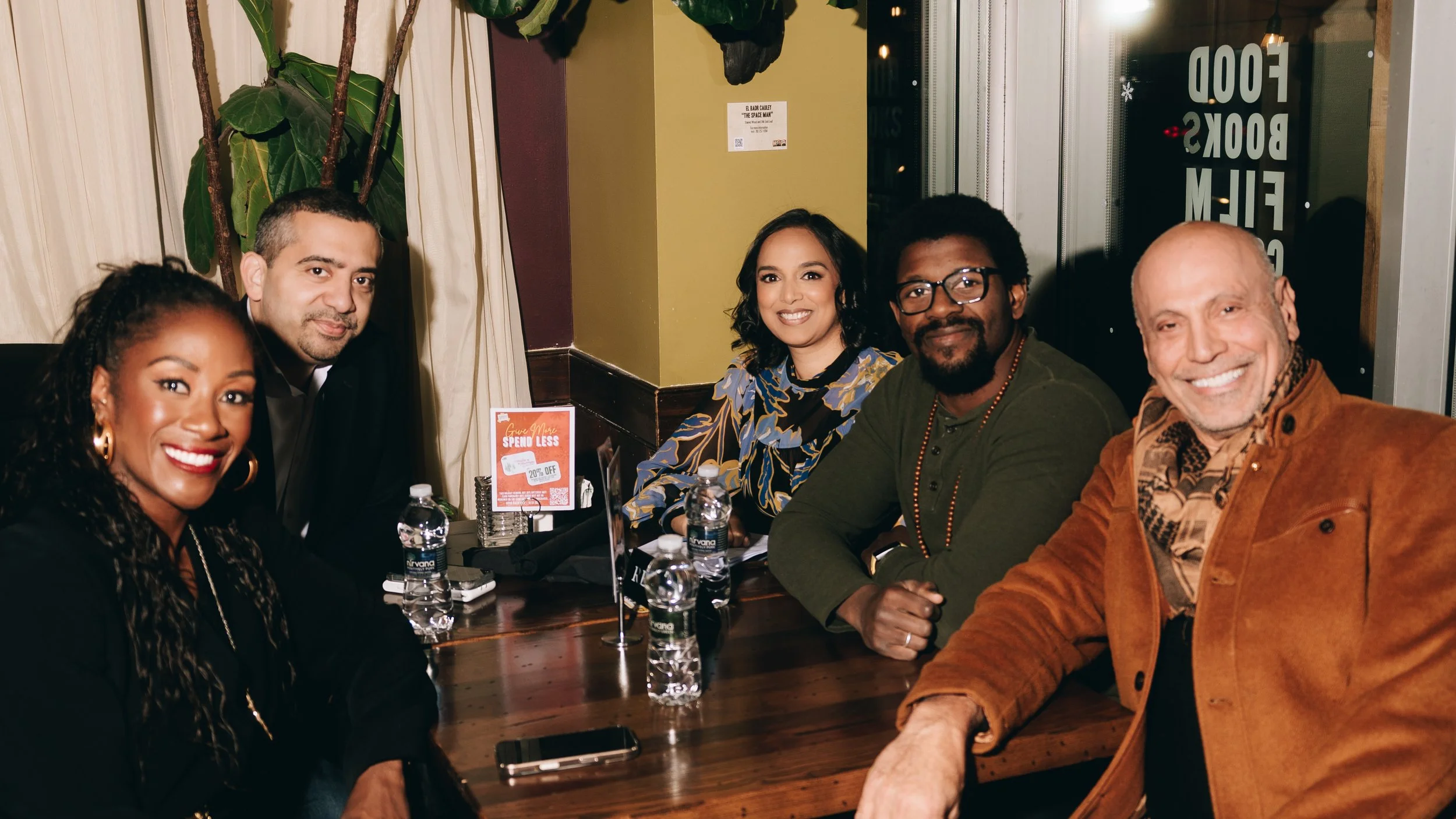Five people sitting at a restaurant table, smiling and posing for the photo, with water bottles and a small red sign on the table.