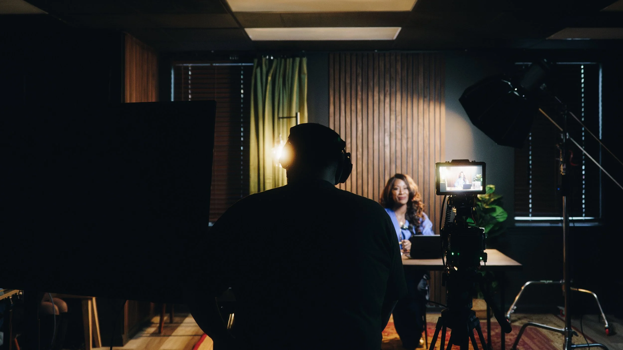 A woman in a blue blazer is being filmed in a recording studio or interview room by a man wearing headphones and a black shirt. The scene is lit by studio lighting, with wooden wall panels and green curtains in the background.