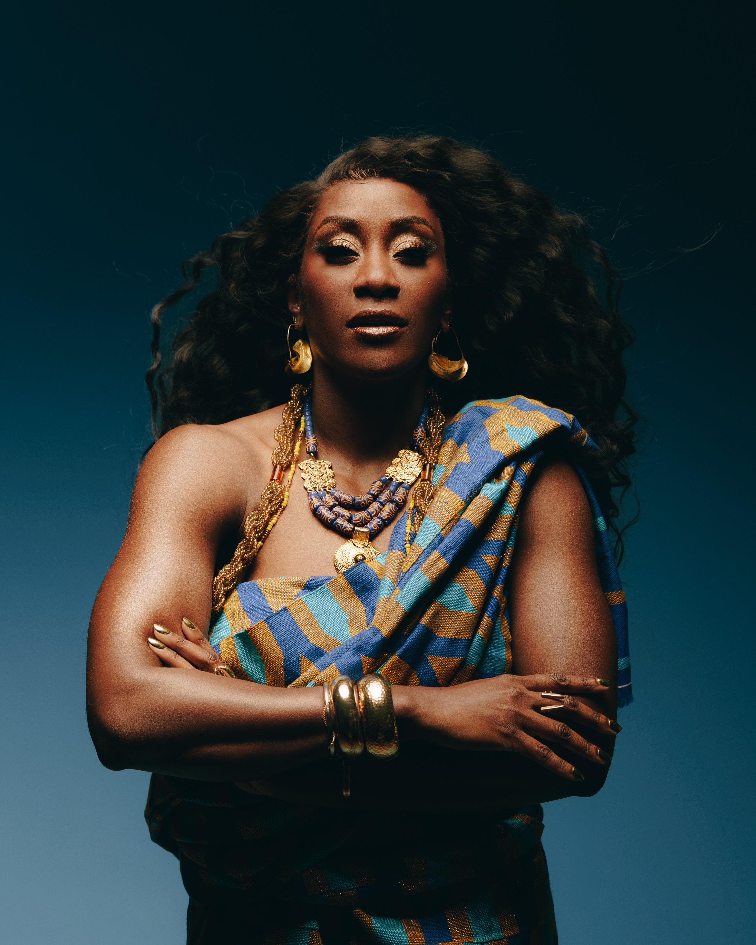 A woman with dark curly hair wearing traditional African jewelry and a colorful patterned fabric draped over her shoulder, standing with arms crossed against a dark background.