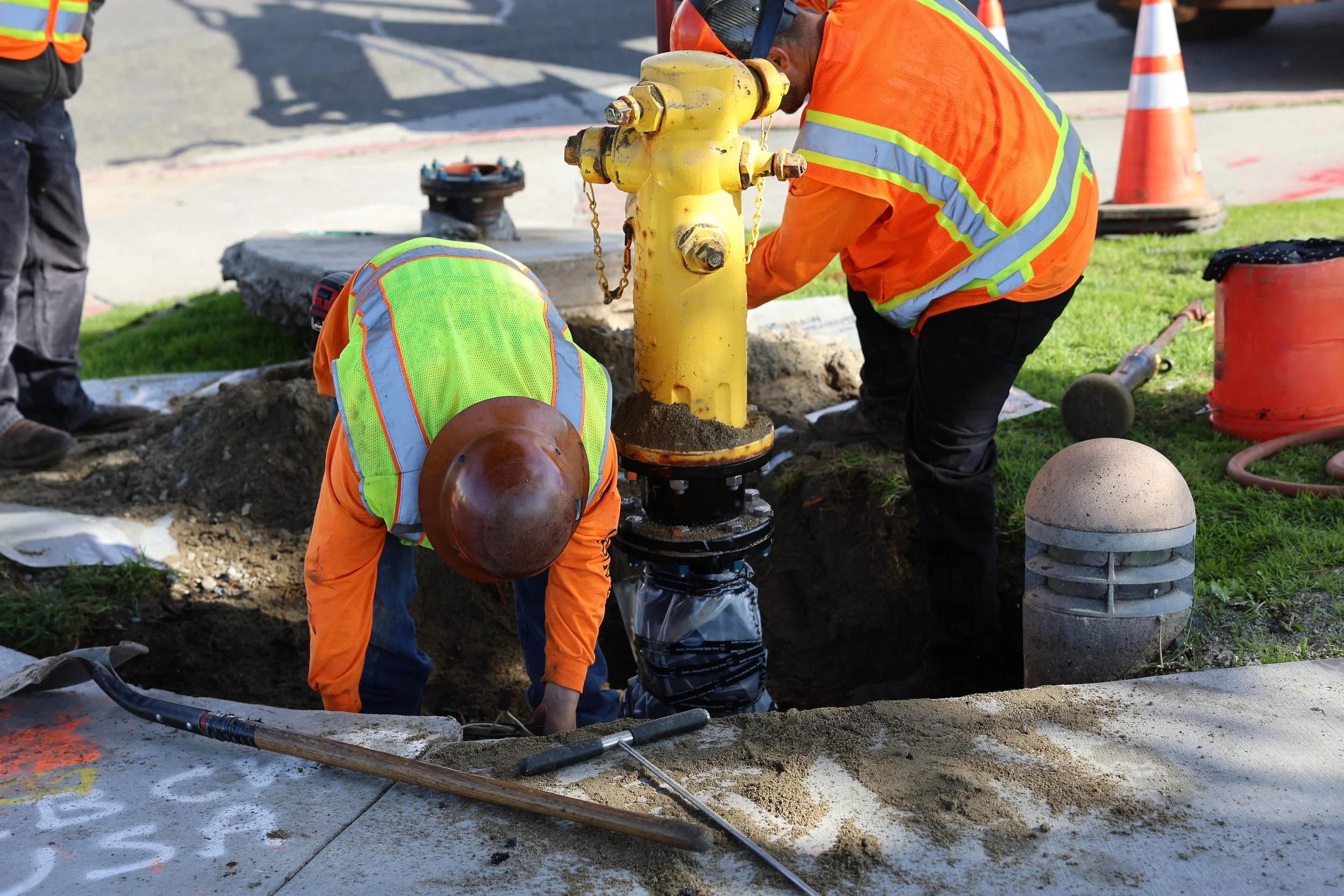 Two workers in orange safety vests and helmets working on underground infrastructure, installing or repairing a fire hydrant with a yellow valve.