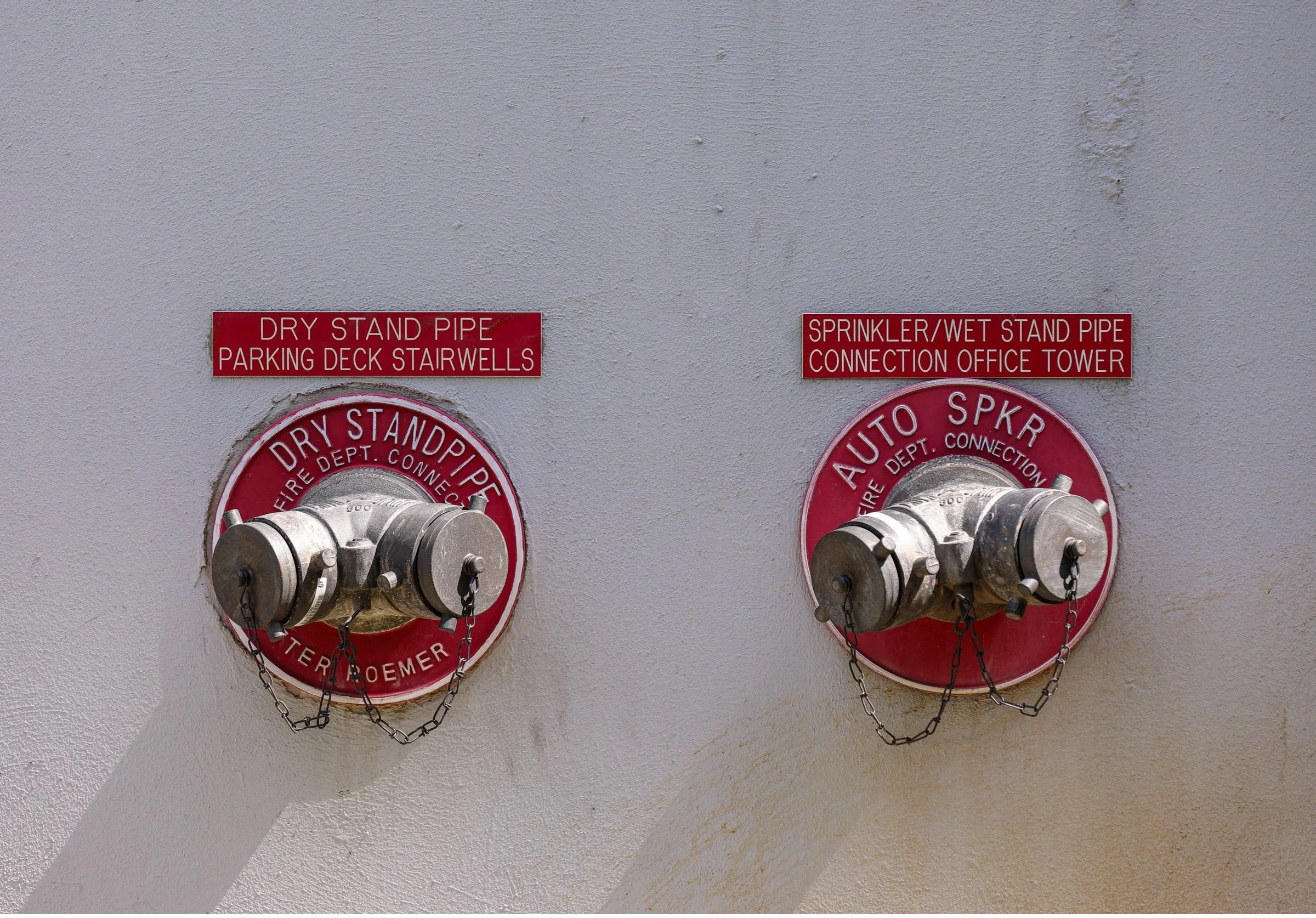 Two fire department sprinkler and standpipe connection valves mounted on a wall, with red informational signs above. The left sign reads 'Dry Stand Pipe Parking Deck Stairwells,' and the right sign reads 'Sprinkler/Wet Stand Pipe Connection Office Tower.' The valves are metallic, with chains attached.