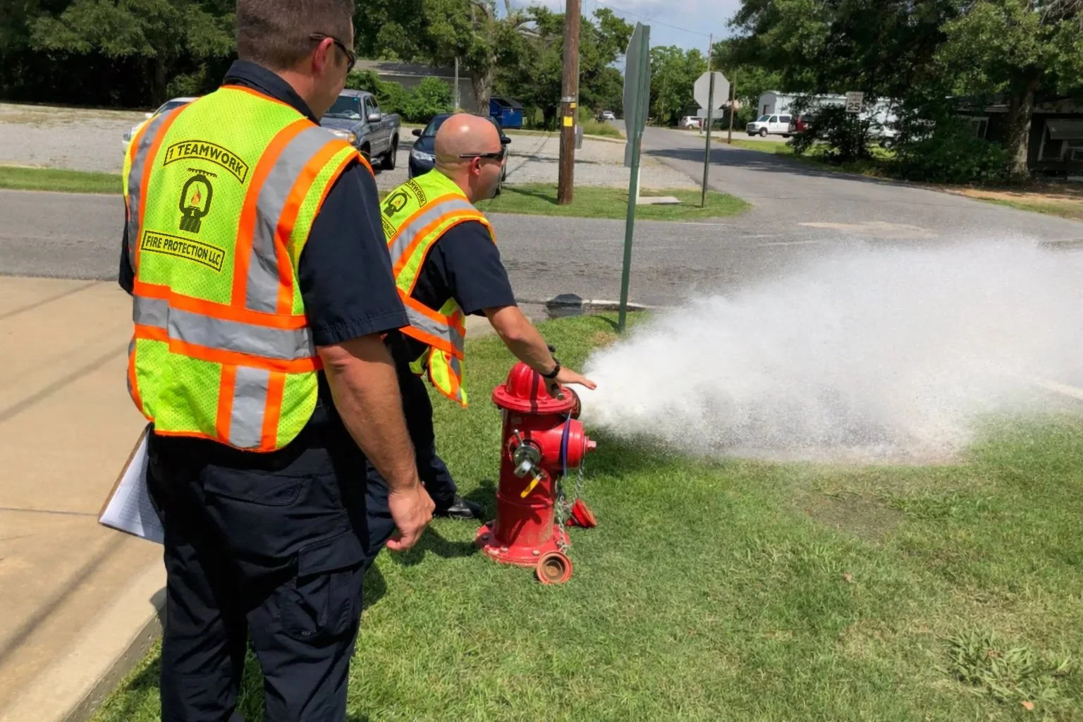 Two firefighters in yellow safety vests with '1 Teamwork' and 'Fire Protection LLC' printed on the back, inspecting a fire hydrant with water spraying out of it on a grassy area in a neighborhood.