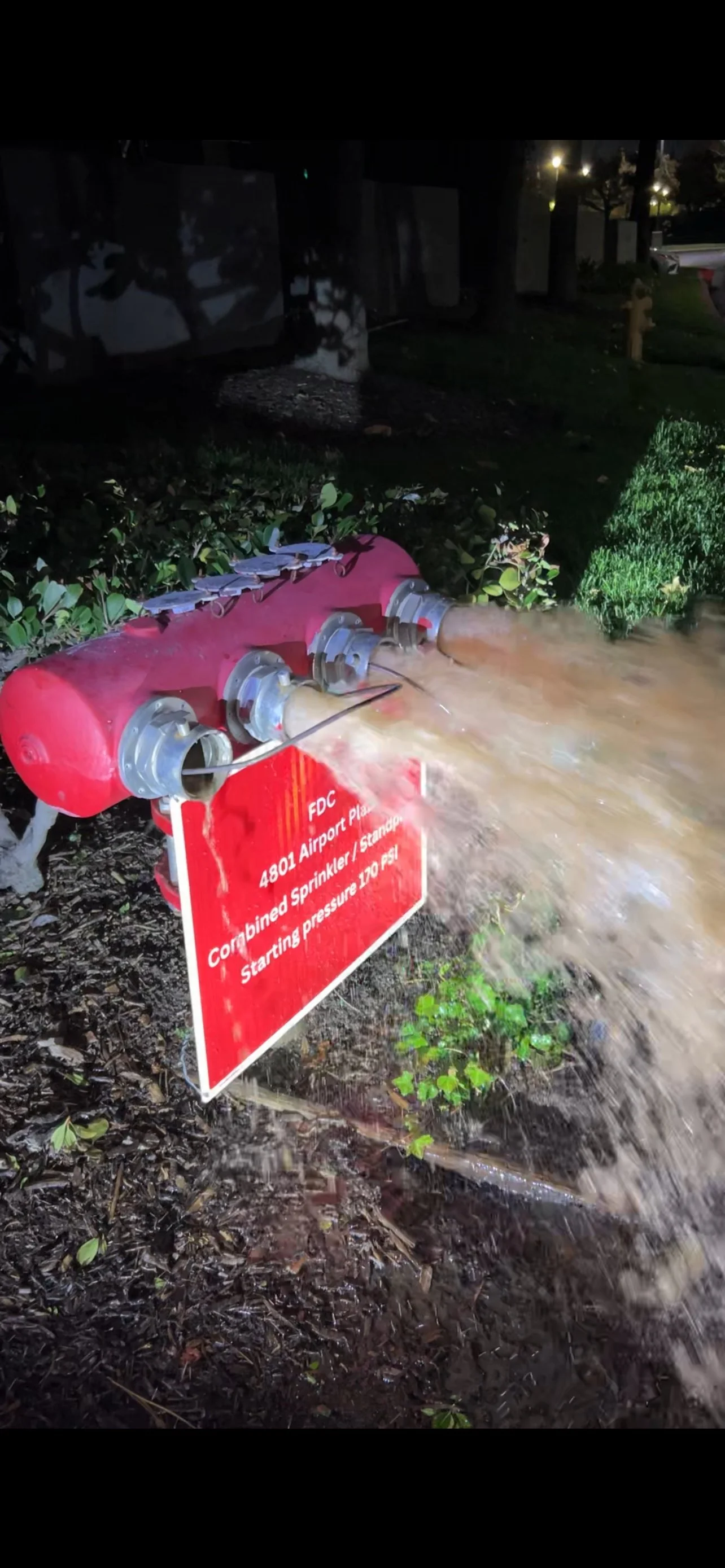 A water pipe leaking or spraying water onto the ground, with a red sign in front of it indicating it is part of an airport's sprinkler system.