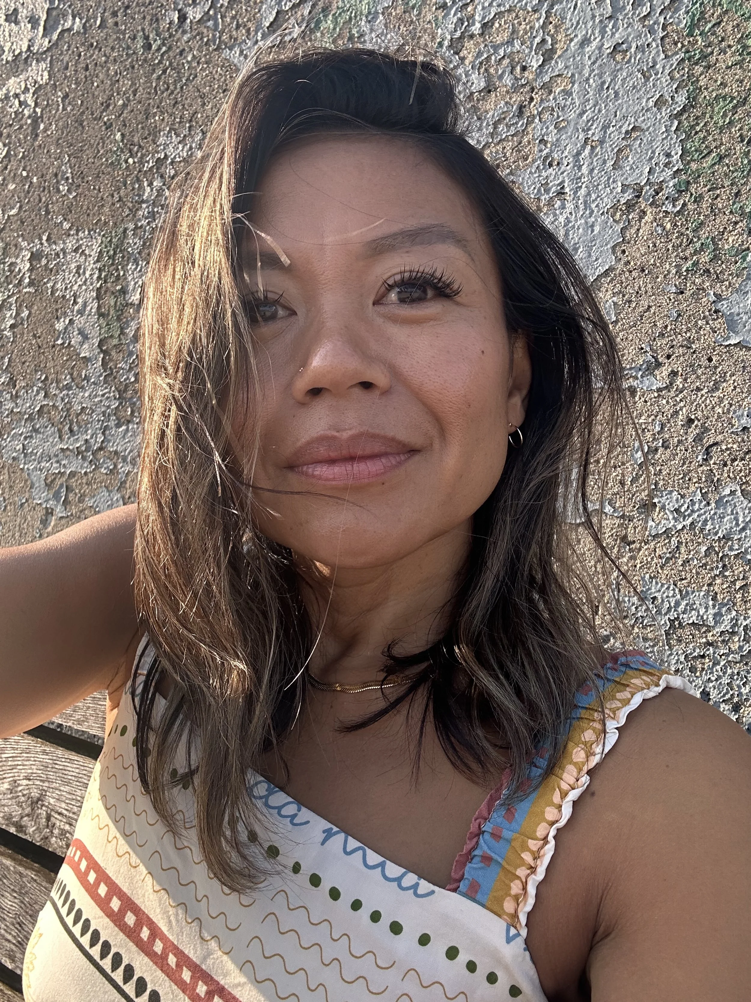 Close-up of a woman with shoulder-length dark hair taking a selfie outdoors against a textured wall.