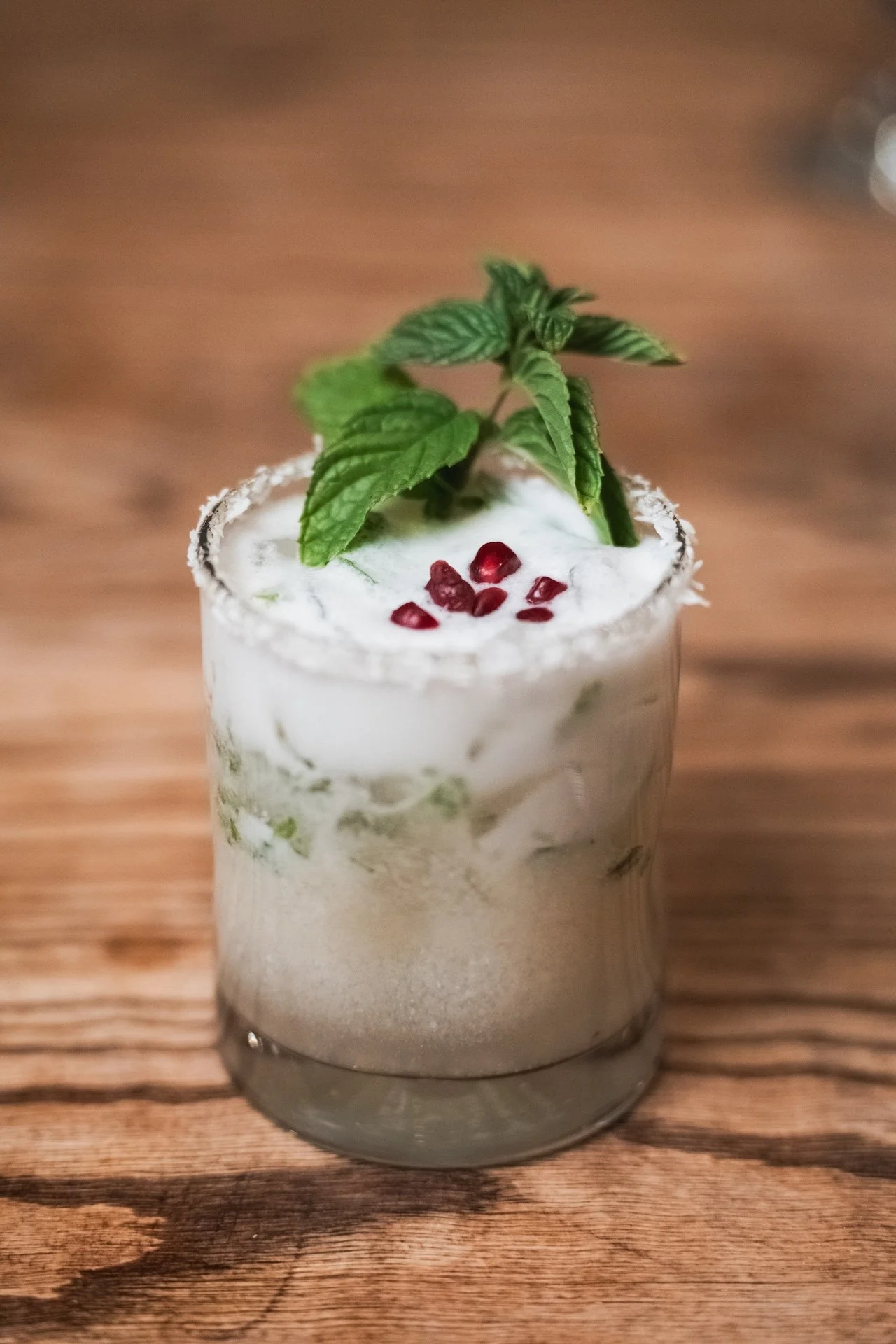 A cocktail in a clear glass garnished with mint leaves, pomegranate seeds, and whipped cream, on a wooden surface. Food Photographer in Kalispell, Montana in the Flathead Valley