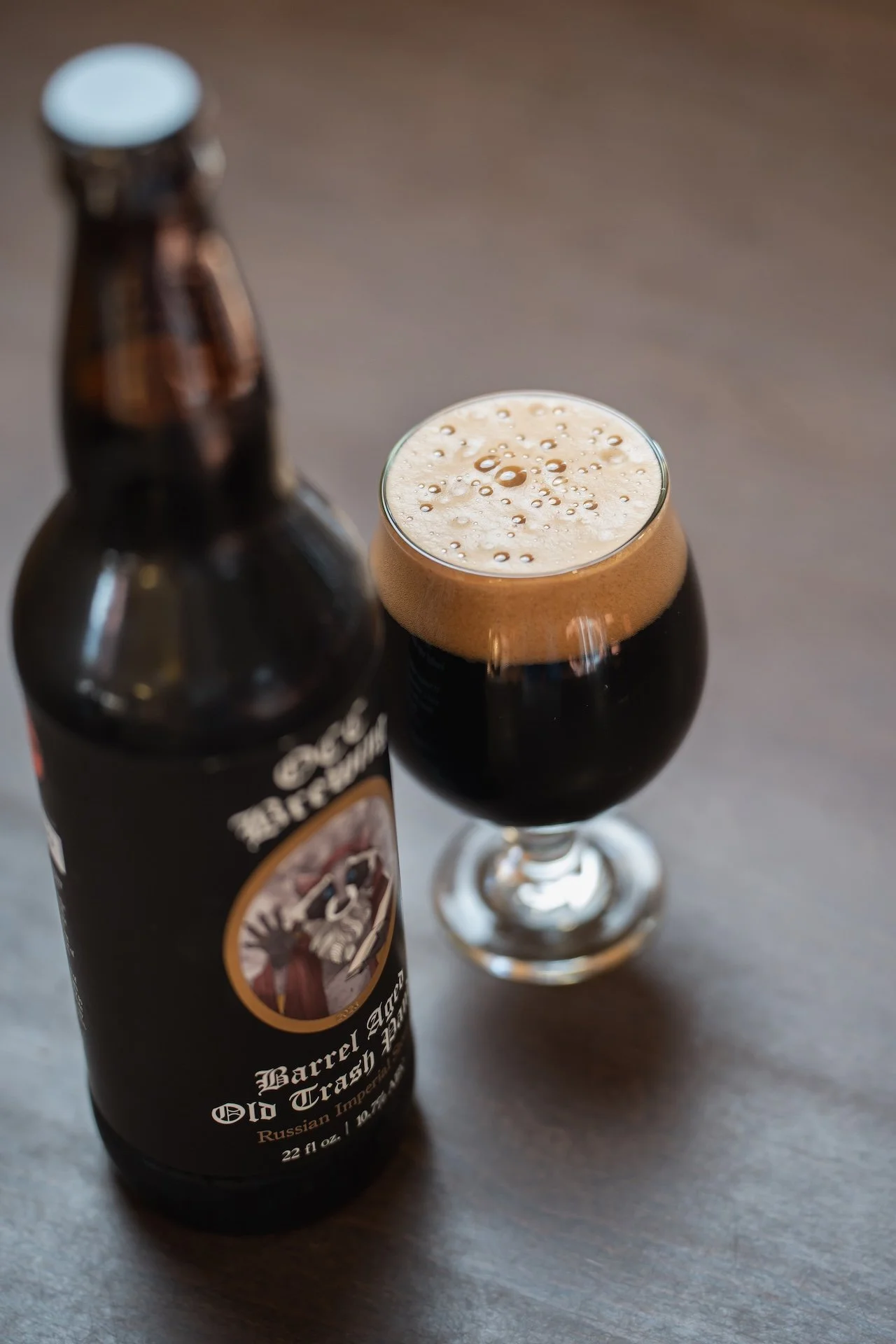 A dark beer with a foamy head in a tulip glass next to a brown bottle labeled "Old Trapper Russian Imperial," set on a wooden surface. Food Photographer in Kalispell, Montana in the Flathead Valley