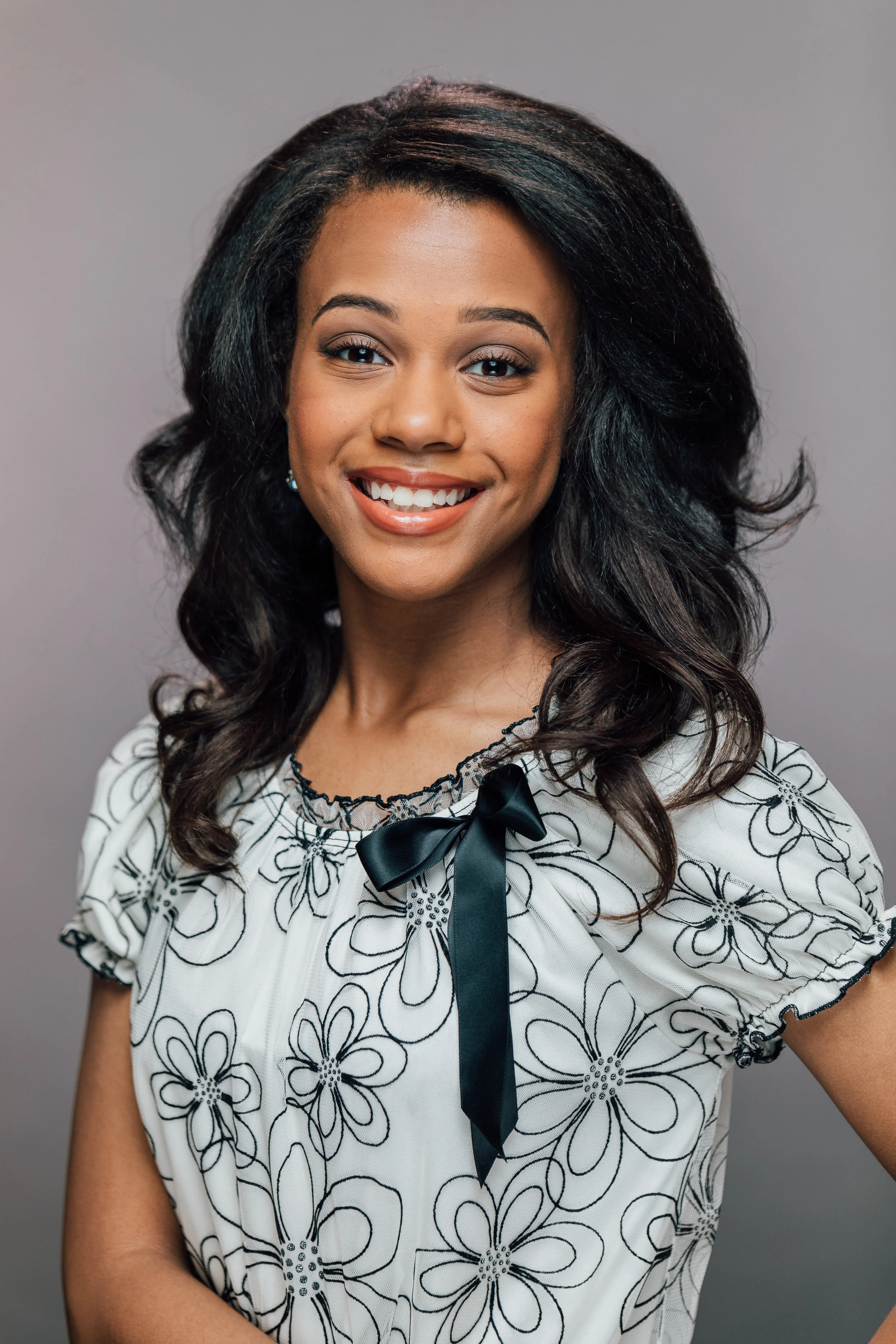 Smiling woman with long dark hair wearing a floral patterned blouse and bow against a gray background. Professional Headshot and Portrait Photographer in Kalispell, Montana in the Flathead Valley.