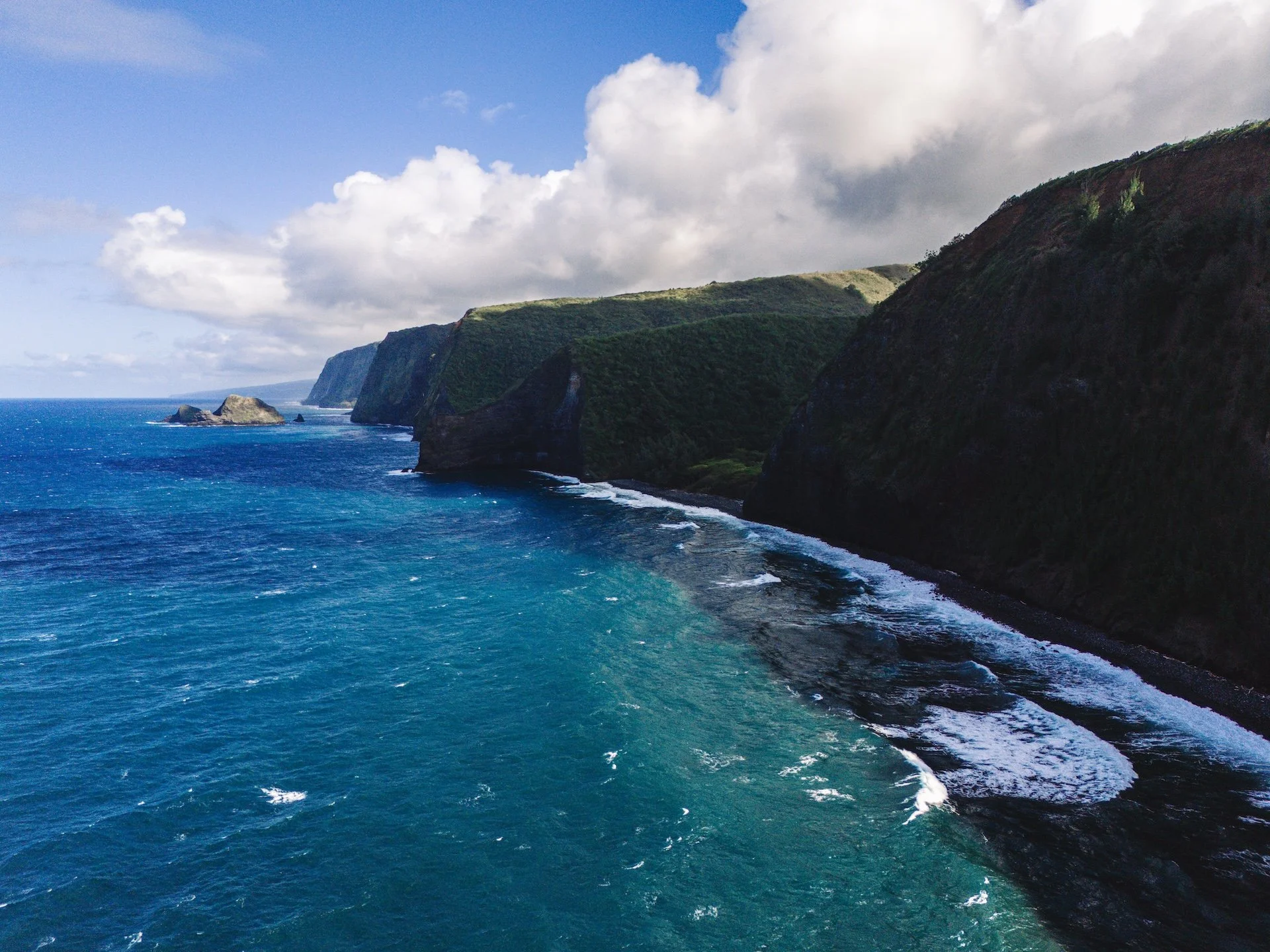 Aerial view of rocky cliffs along a blue ocean, with waves crashing onto the dark shoreline, under a partly cloudy sky.