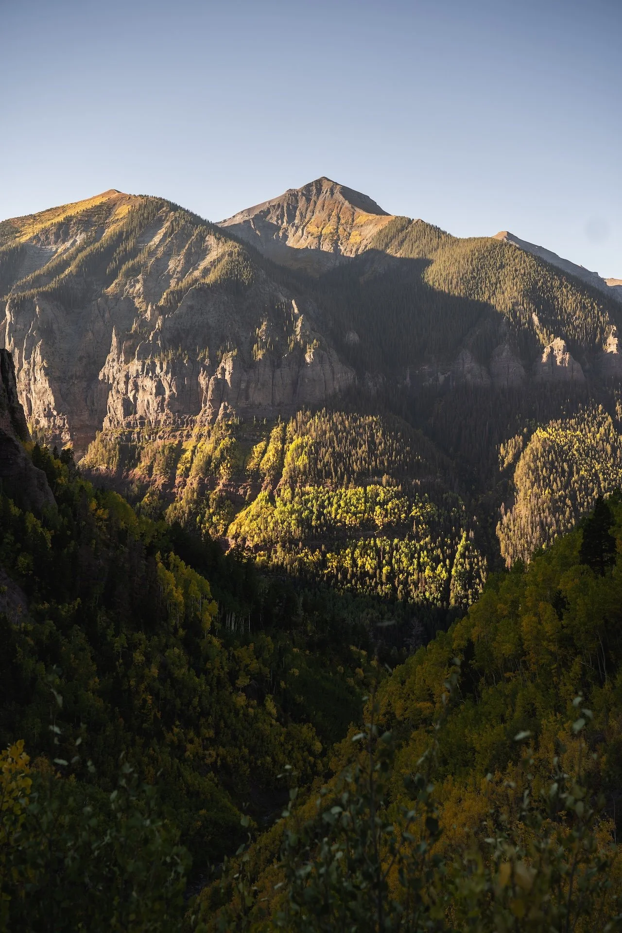 A scenic mountain landscape with rugged peaks, lush green forests, and clear sky.