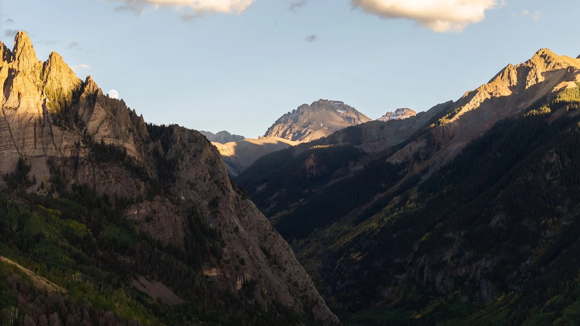 Mountain landscape with rugged peaks, forested slopes, and a partly cloudy sky