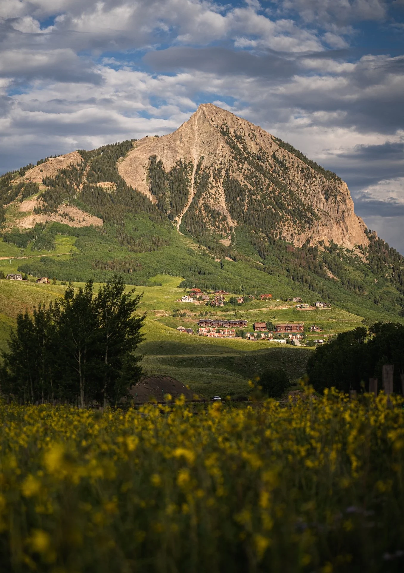 A scenic landscape with a large mountain, a partly cloudy sky, and a small town at the base surrounded by green fields and trees.