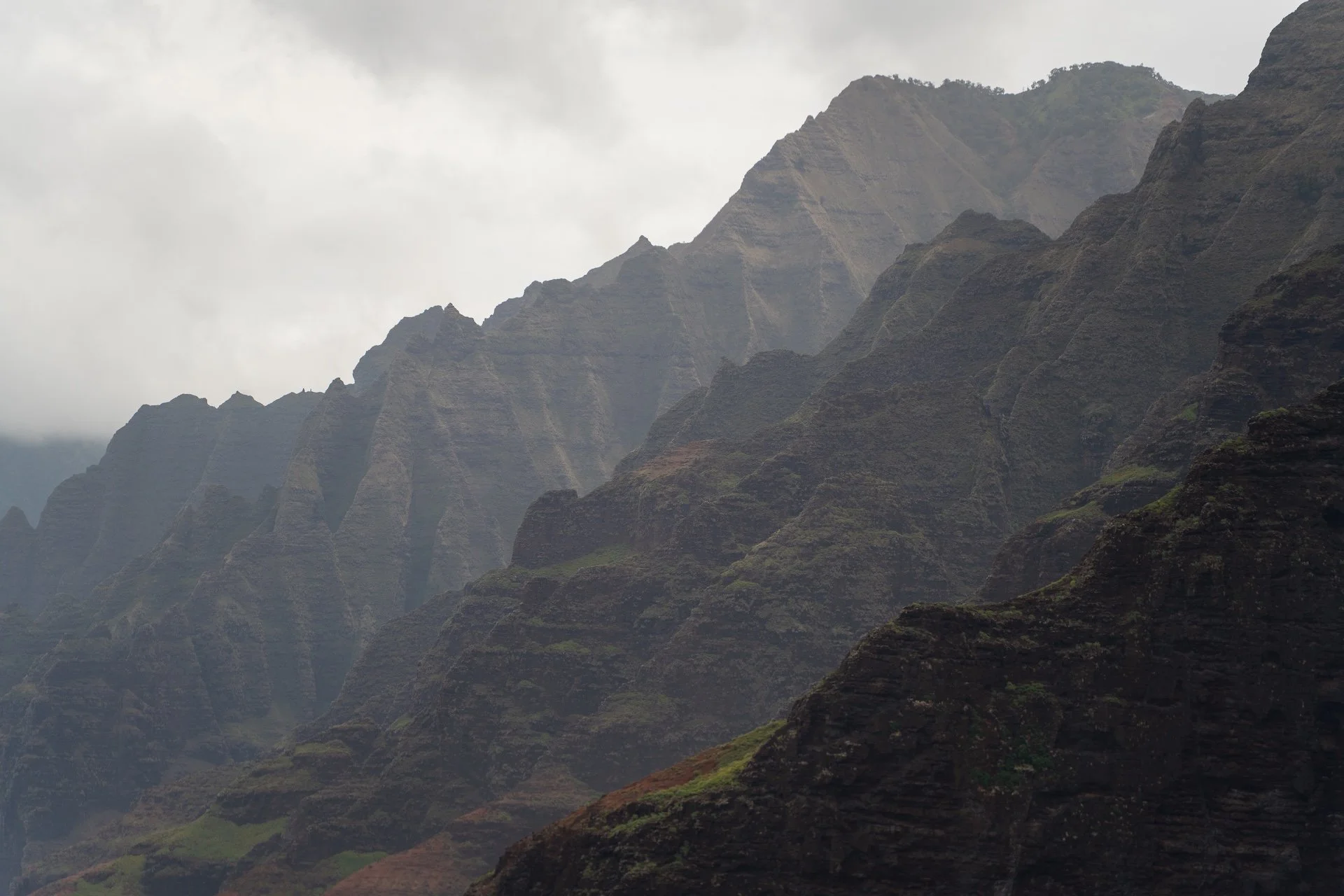 A misty mountain range with jagged peaks and green vegetation on the slopes under a cloudy sky.