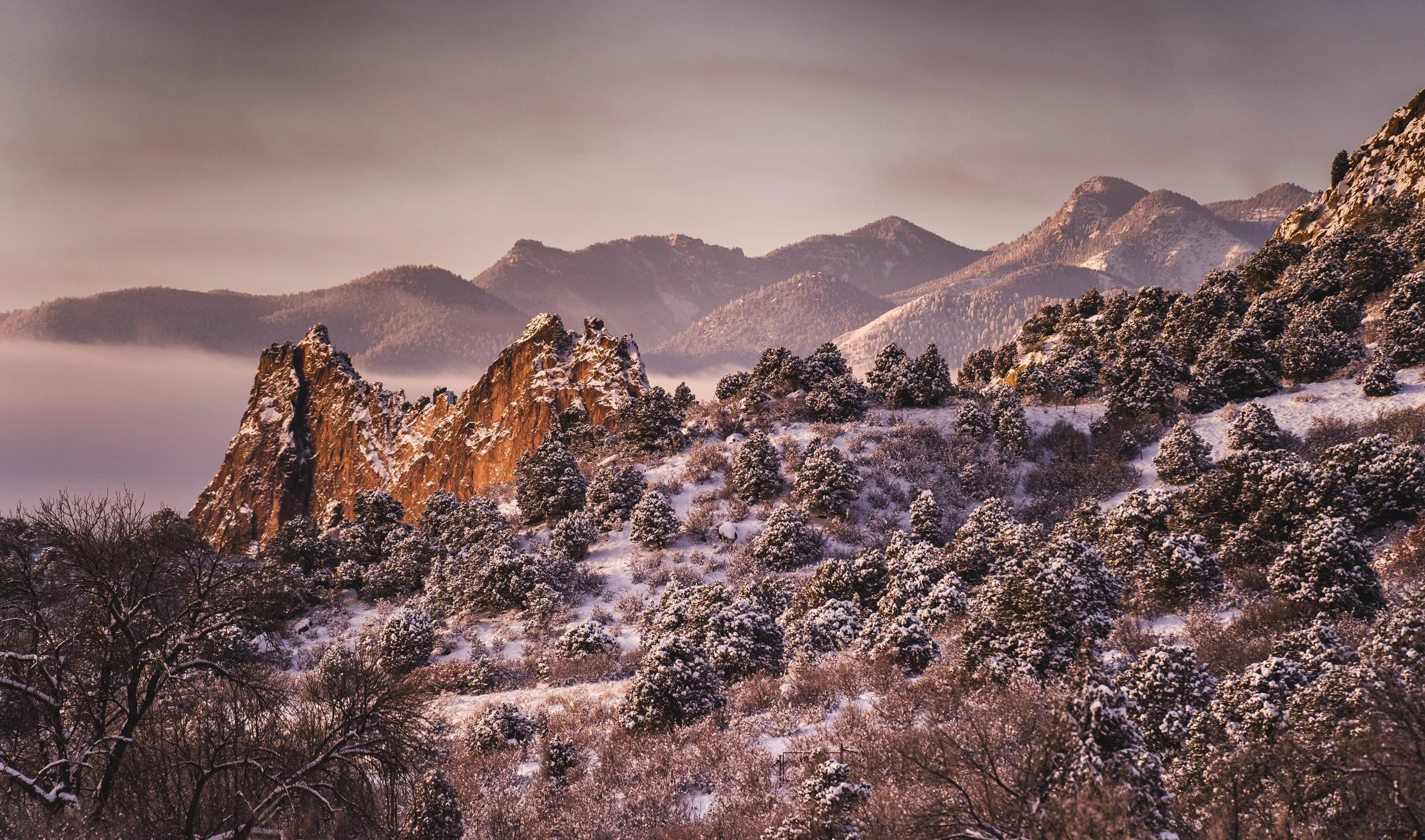 Rocky mountain landscape with snow-covered trees and red rock formations at sunset.
