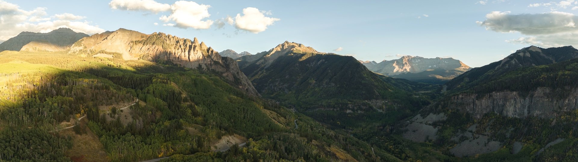 A panoramic view of a mountain valley with lush green forests, rugged mountain peaks, and a winding road running through the valley. The sky is partly cloudy, with sunlight illuminating parts of the mountains.