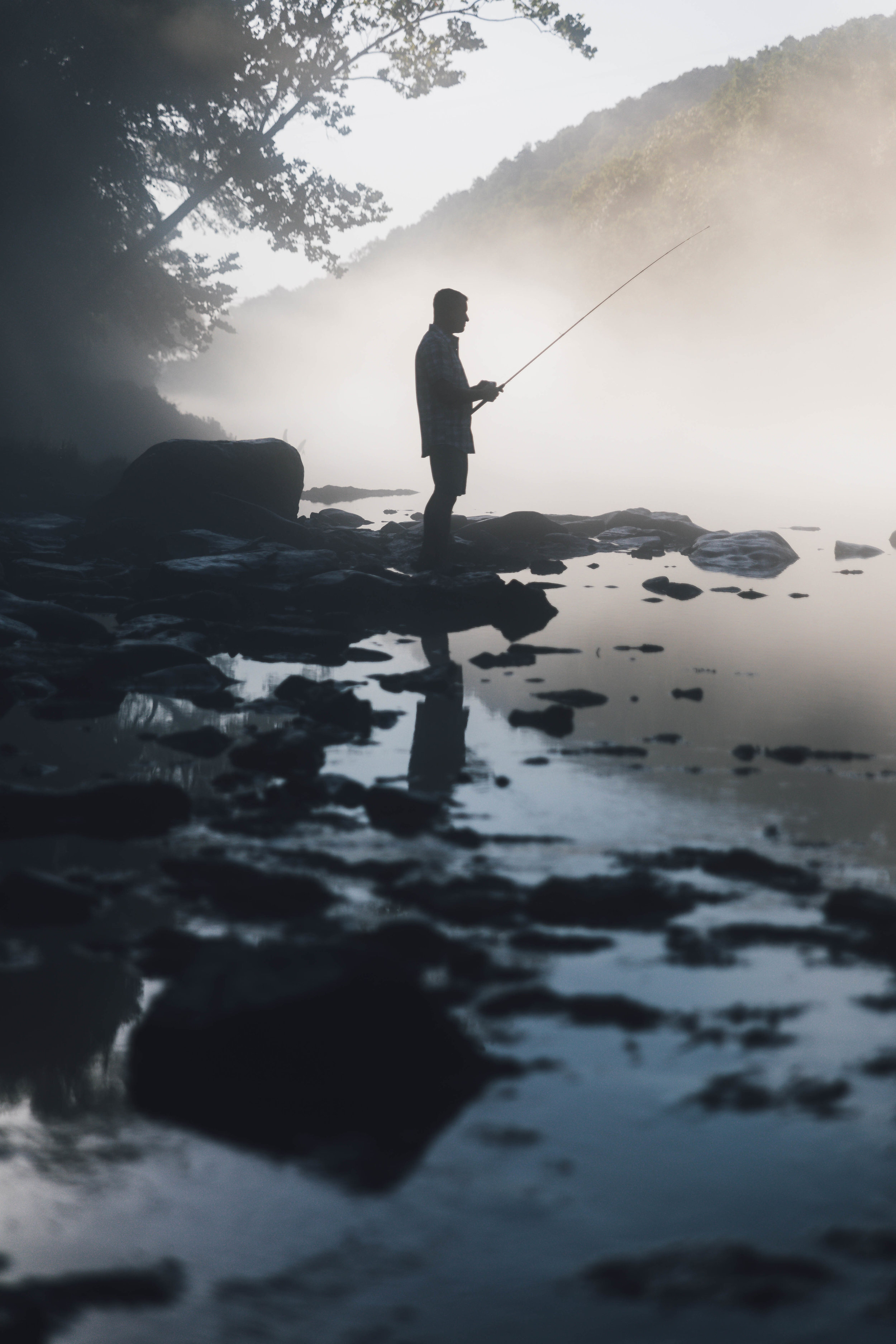 Silhouette of a person fishing by a foggy riverbank, surrounded by rocks and trees, with mist in the background.