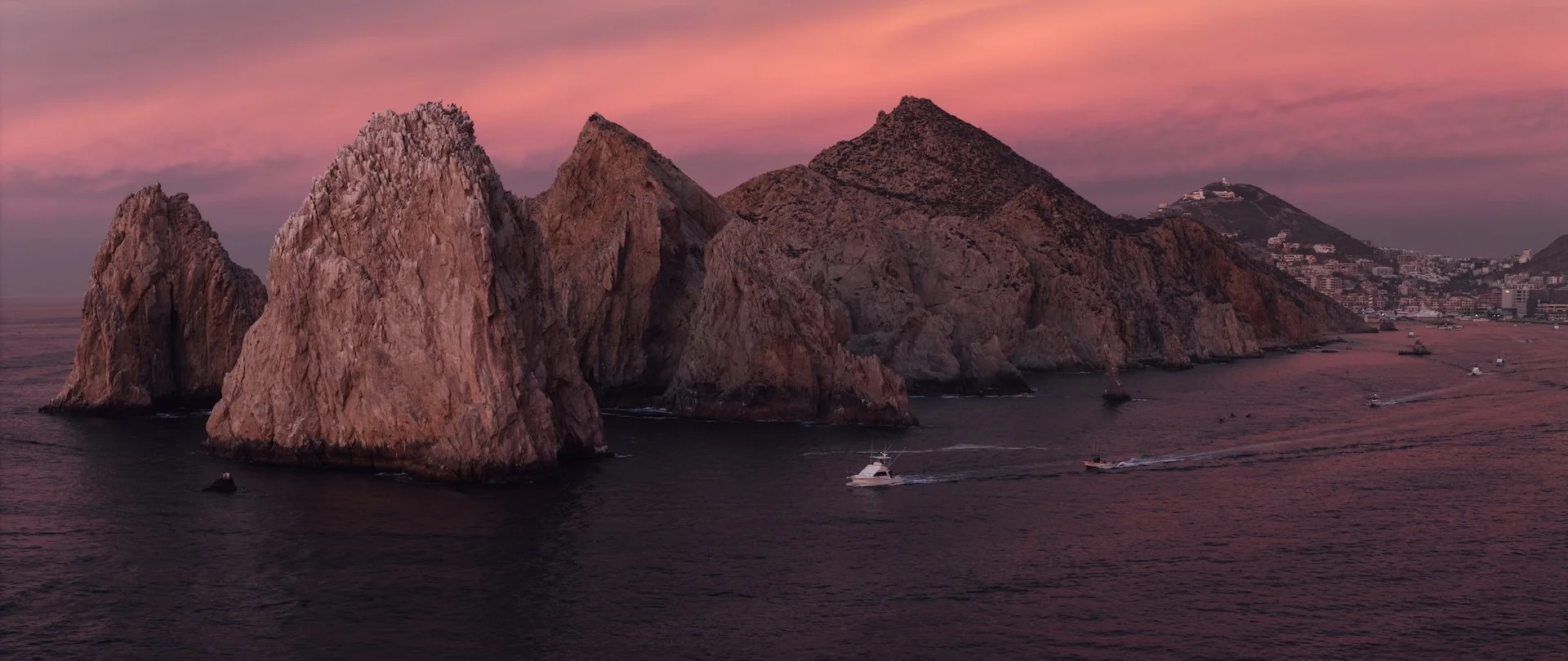 Large rocky islands off the coast during sunset with pink and purple sky, small boats on dark water, a distant city on the hillside.