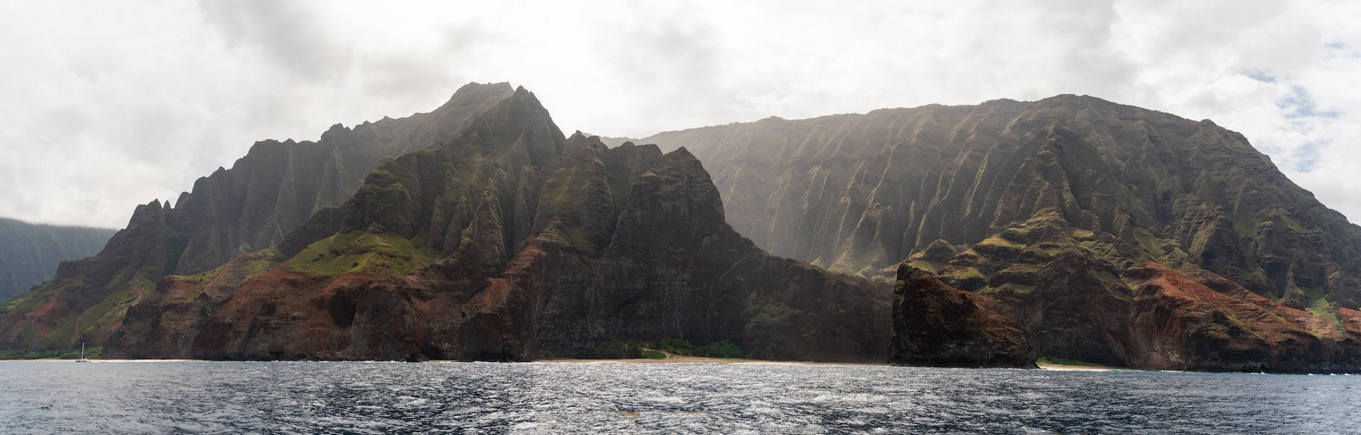 Scenic view of rugged mountains with steep, green slopes rising from the ocean under cloudy sky.