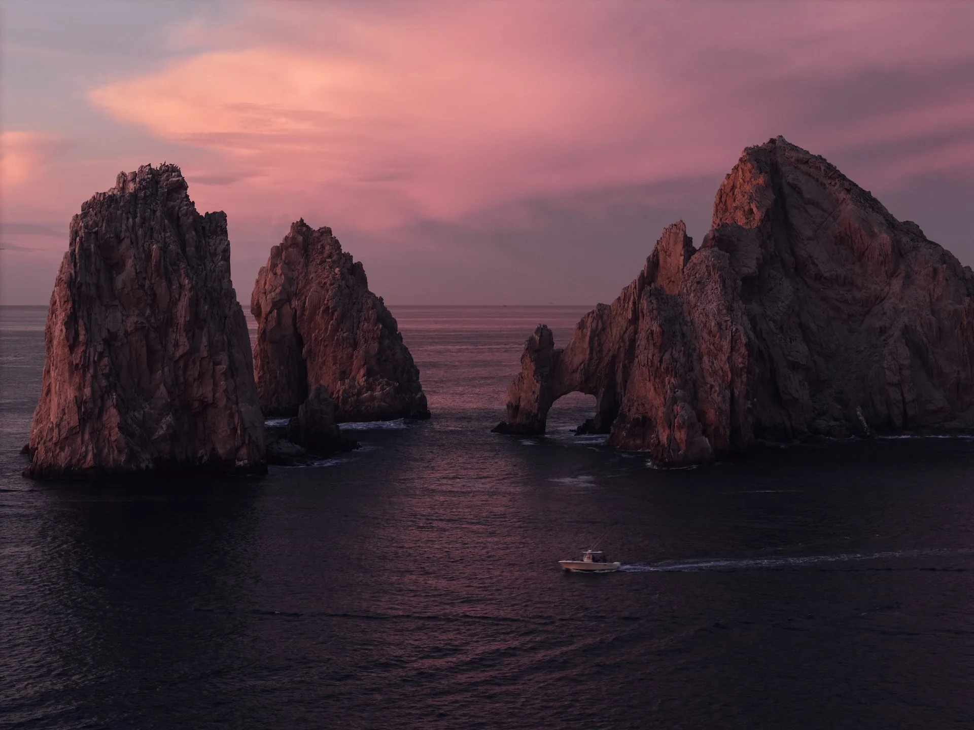 Sunset over sea stacks, with pink and purple sky and a boat leaving a wake in dark water.