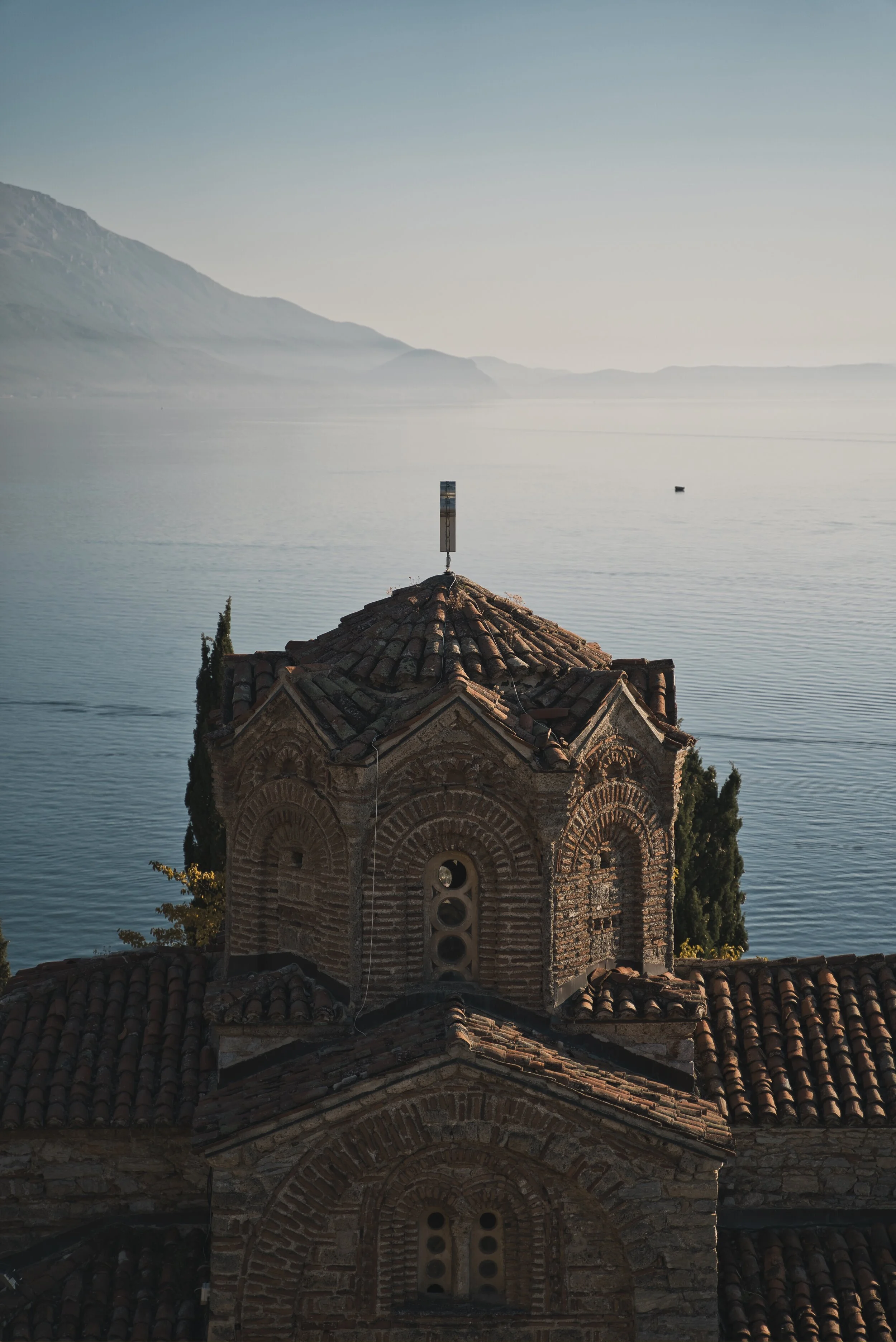 Scenic view of old brick building with arched windows and tiled roof by a calm lake, with mountains in the background.