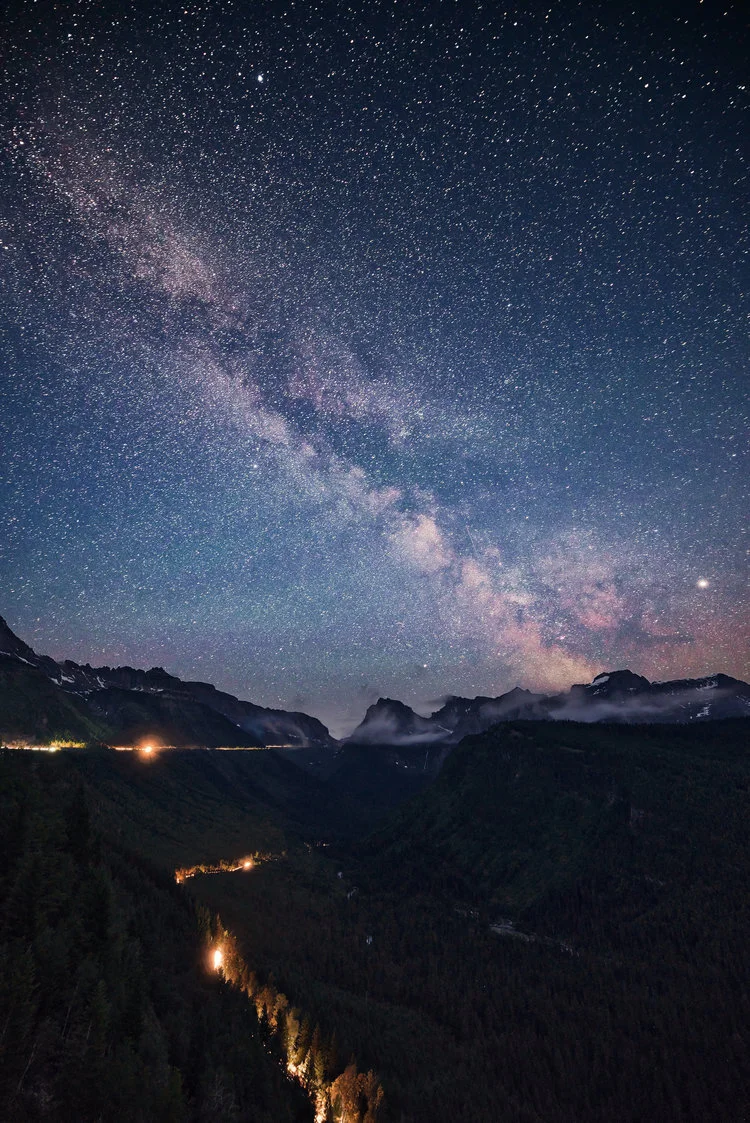Night sky with the Milky Way over a mountain valley, forested hills with scattered lights.