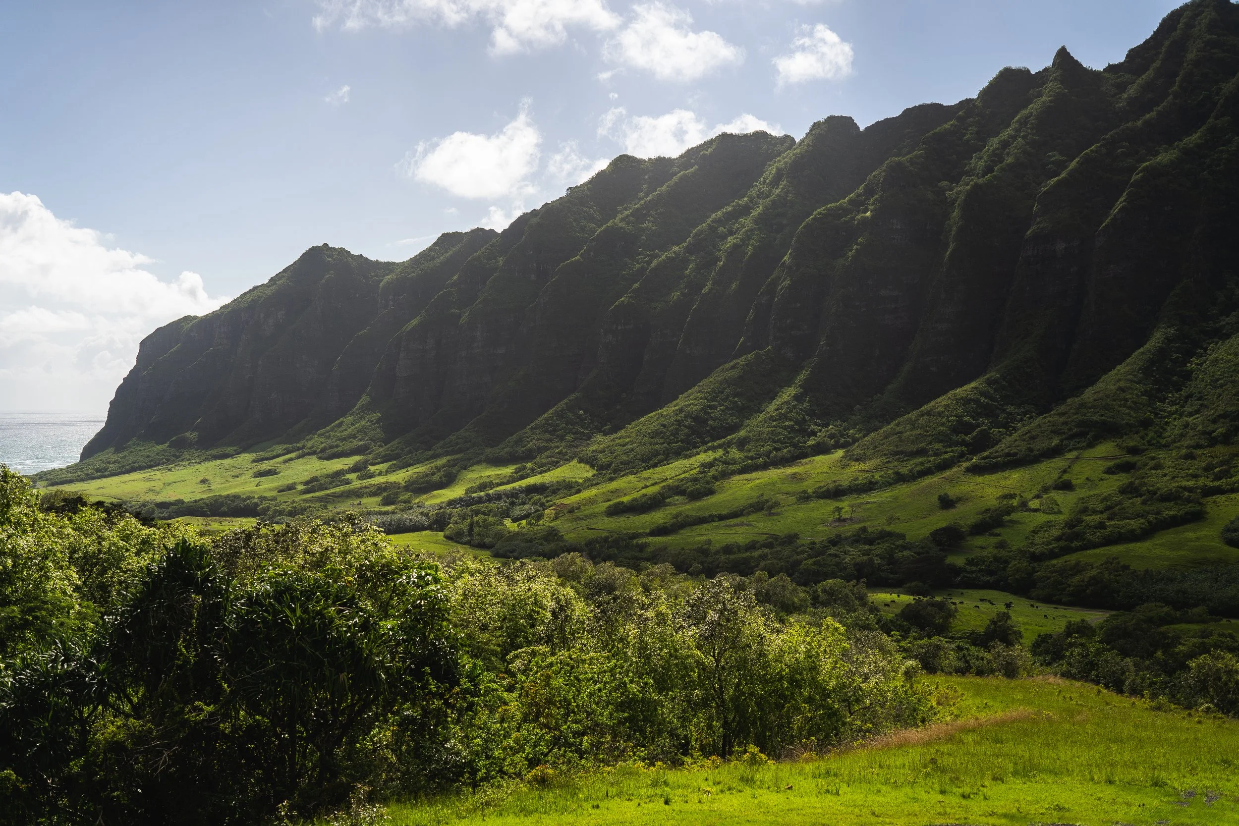 Lush green mountains with steep cliffs and a green valley in front, under a partly cloudy sky.