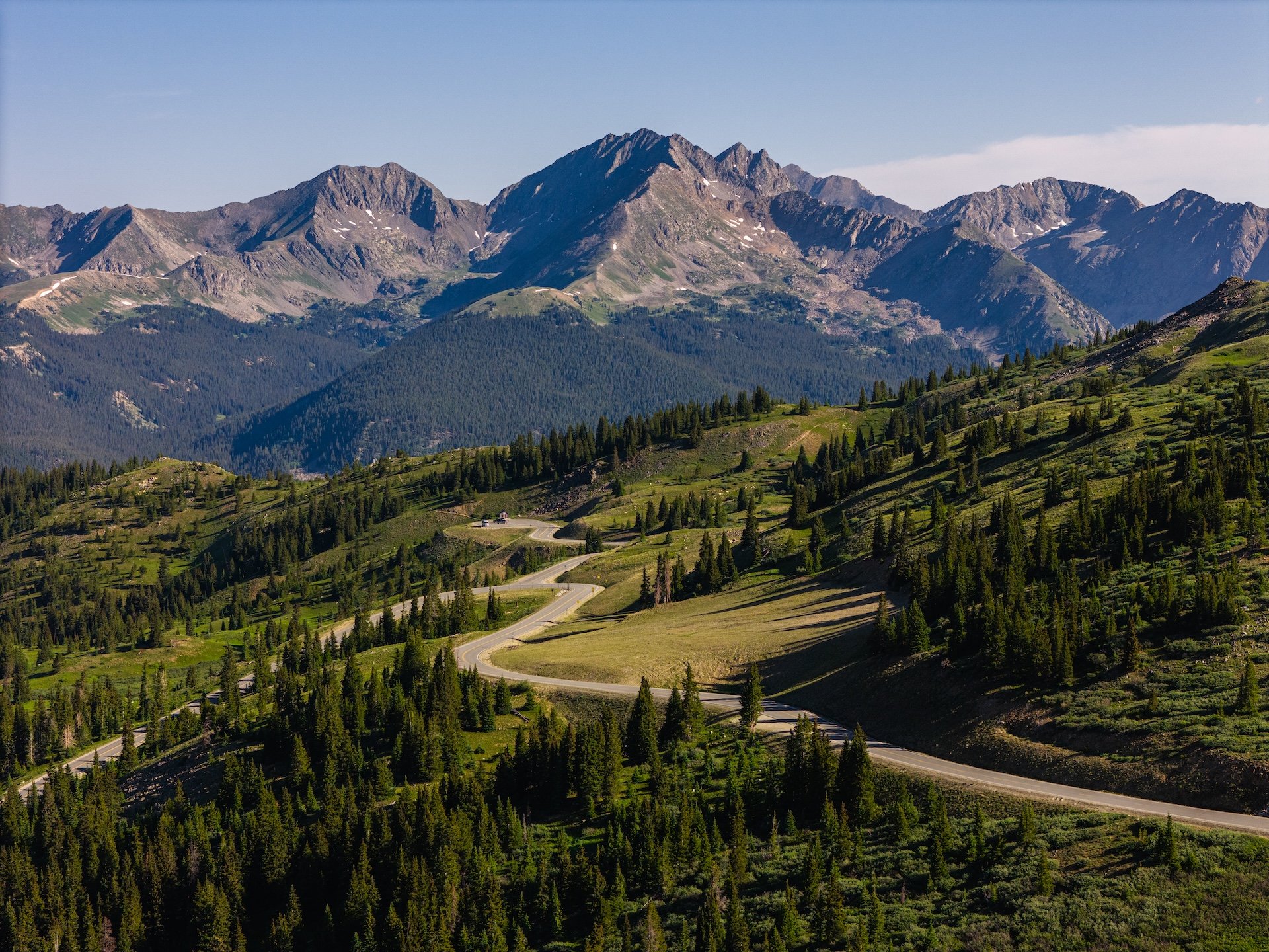 Scenic view of winding mountain road through lush green hills with tall pine trees, with rugged mountains in the background under a blue sky.